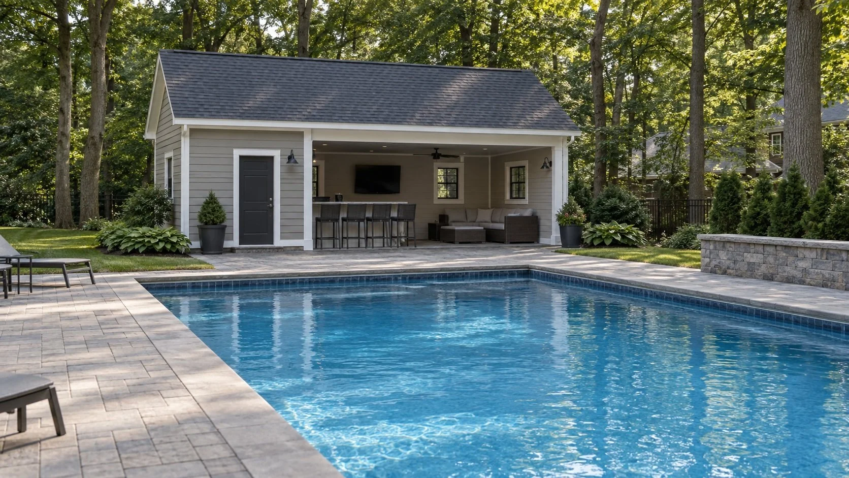 Gray pool house with covered bar, lounge seating, and TV set beside a rectangular in-ground pool