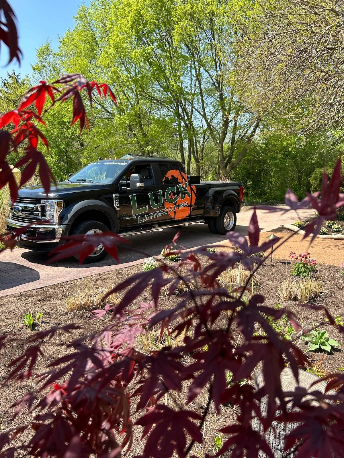 A black pickup truck with a large logo that says "Luck Landscaping" parked on a driveway, surrounded by trees with green leaves and a flower bed with small plants and flowers.