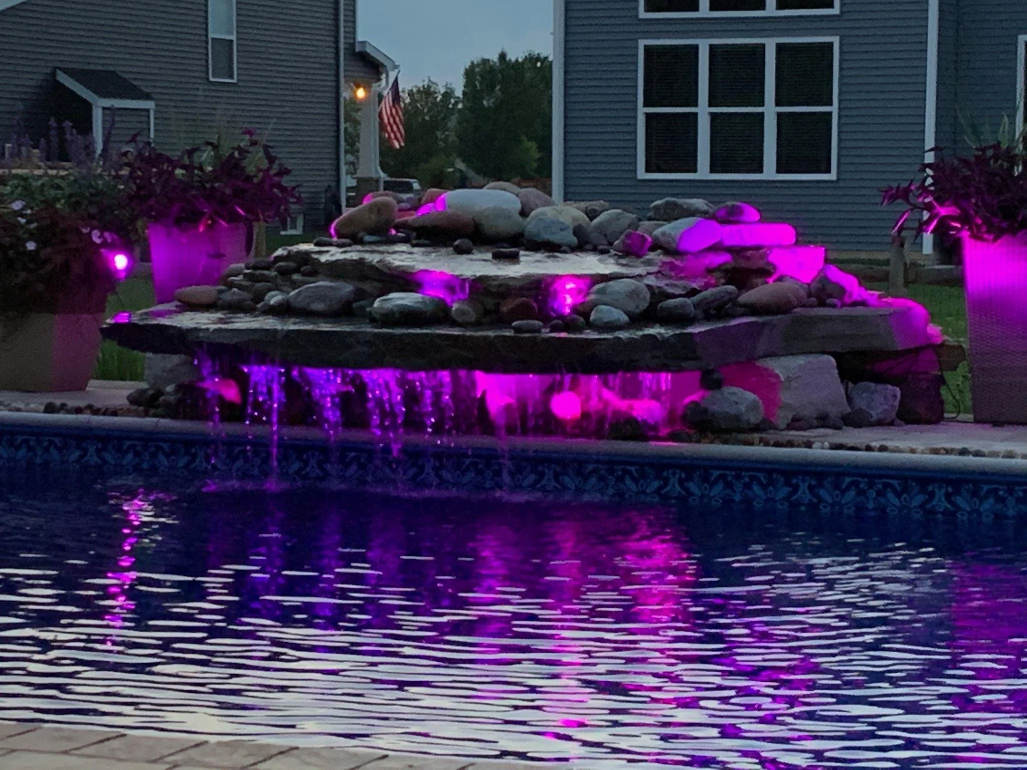 An outdoor fountain with rocks illuminated by purple lights, located in a backyard near a house with gray siding and white-trimmed windows, with potted plants on either side.