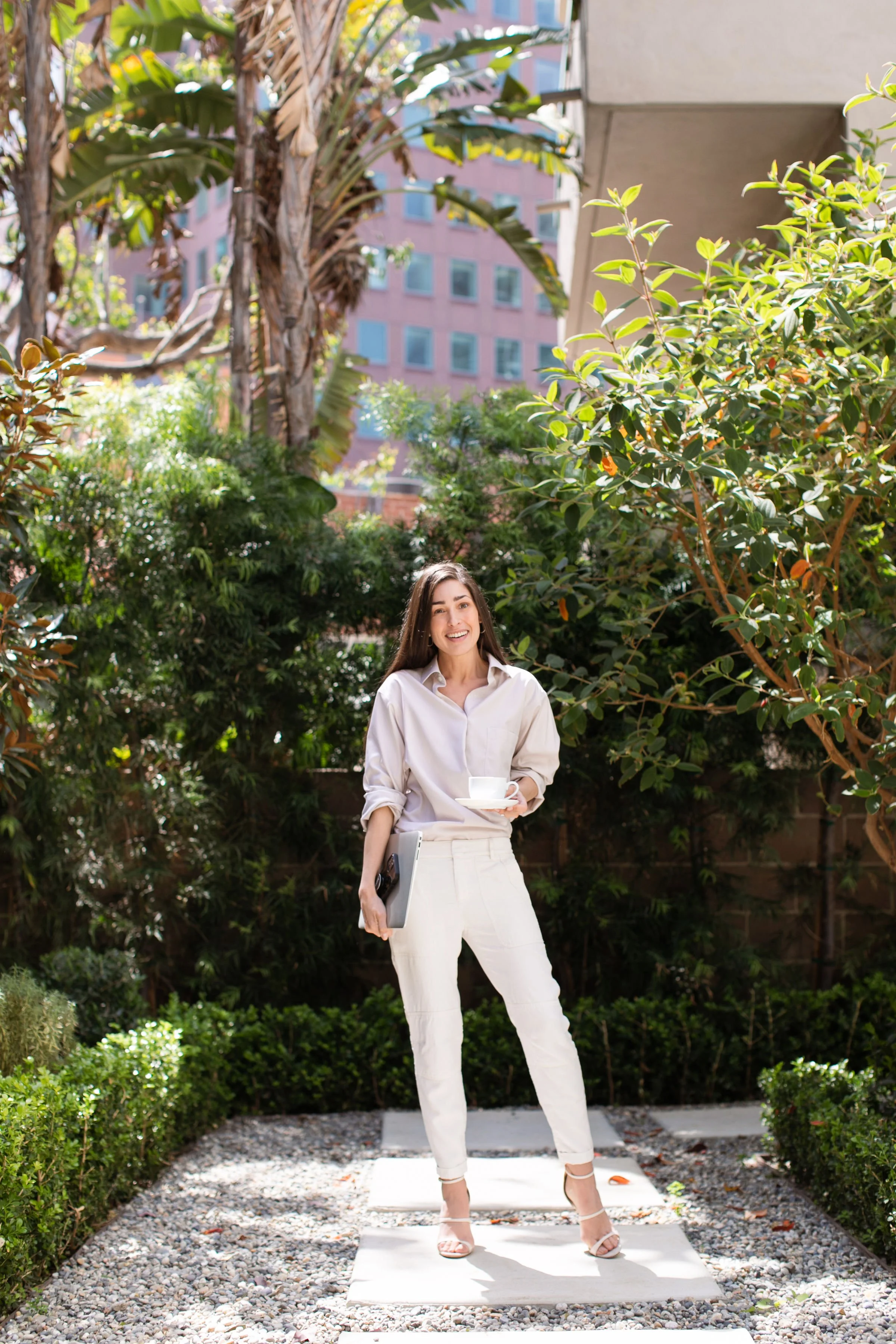 A woman in white pants, a white shirt, and beige high heels stands outdoors on a stone pathway surrounded by lush green bushes and trees, holding a white cup and saucer and a silver laptop, smiling at the camera.