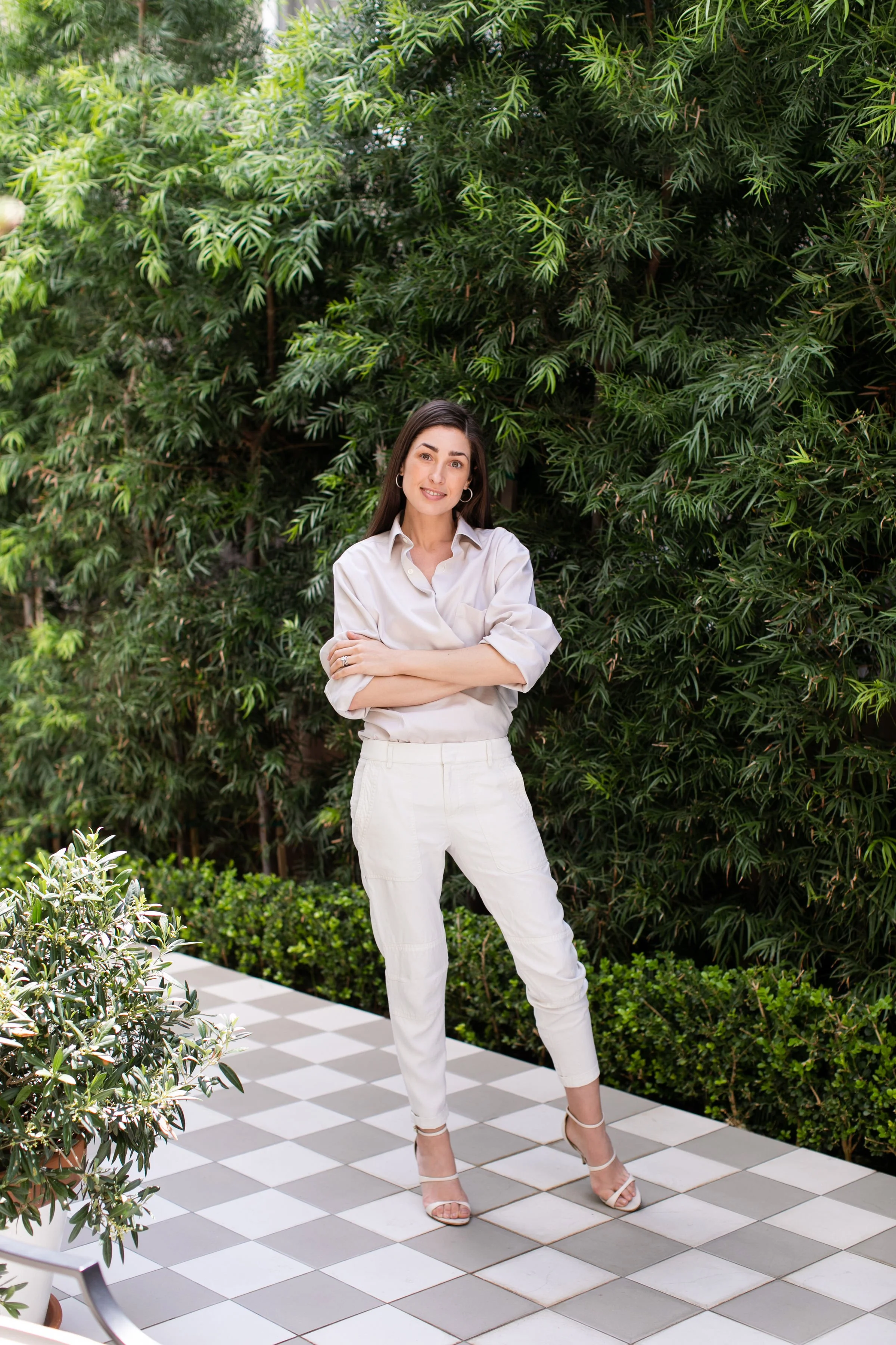 A woman standing on a checkered outdoor patio with green plants and bushes in the background, wearing a white shirt, white pants, and beige high-heeled sandals.