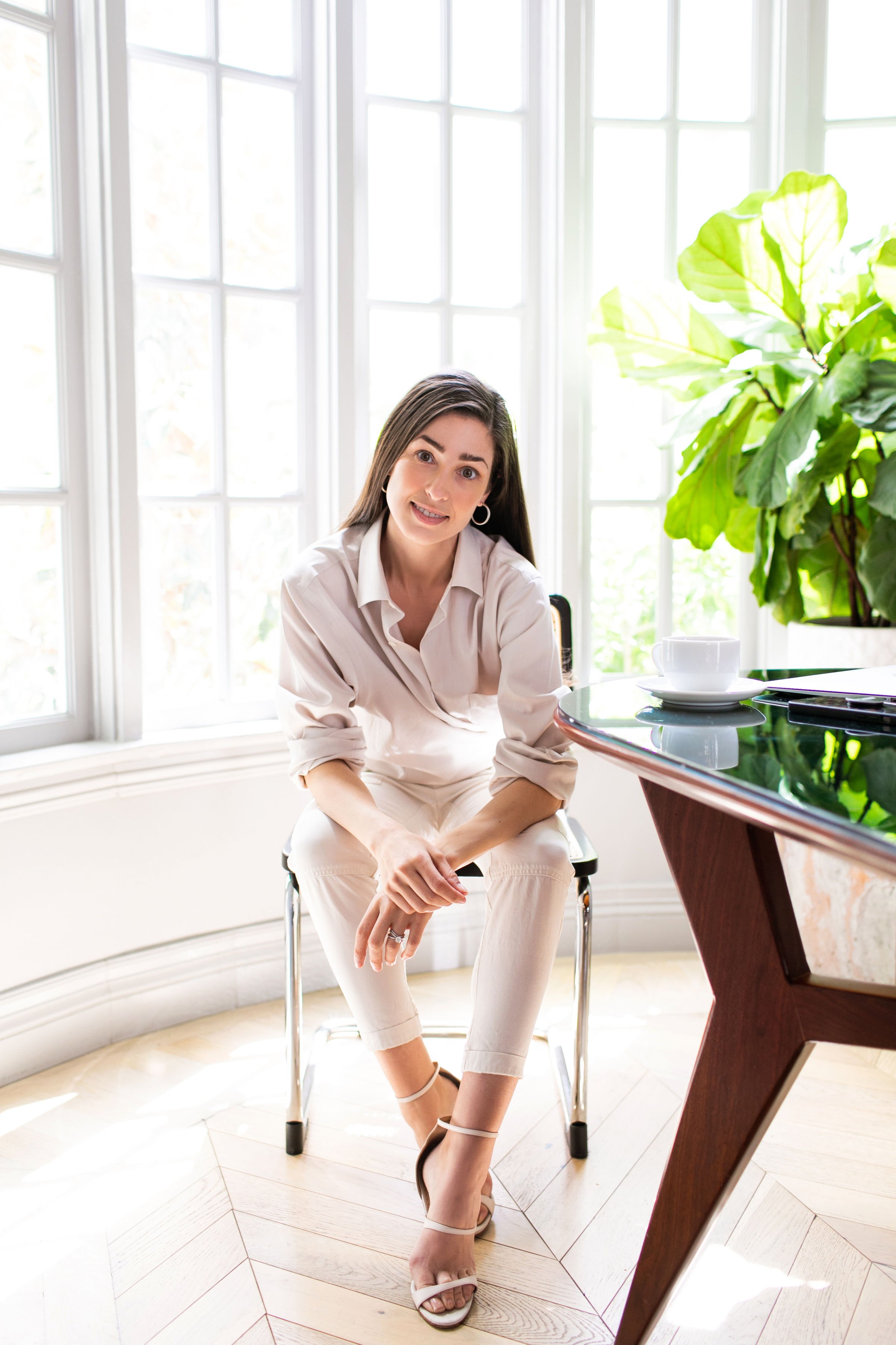 A woman with long dark hair sitting on a chair in a bright room with large windows. She is wearing an off-white outfit, beige heels, and hoop earrings. There is a large green plant and a table with a teacup and saucer nearby.