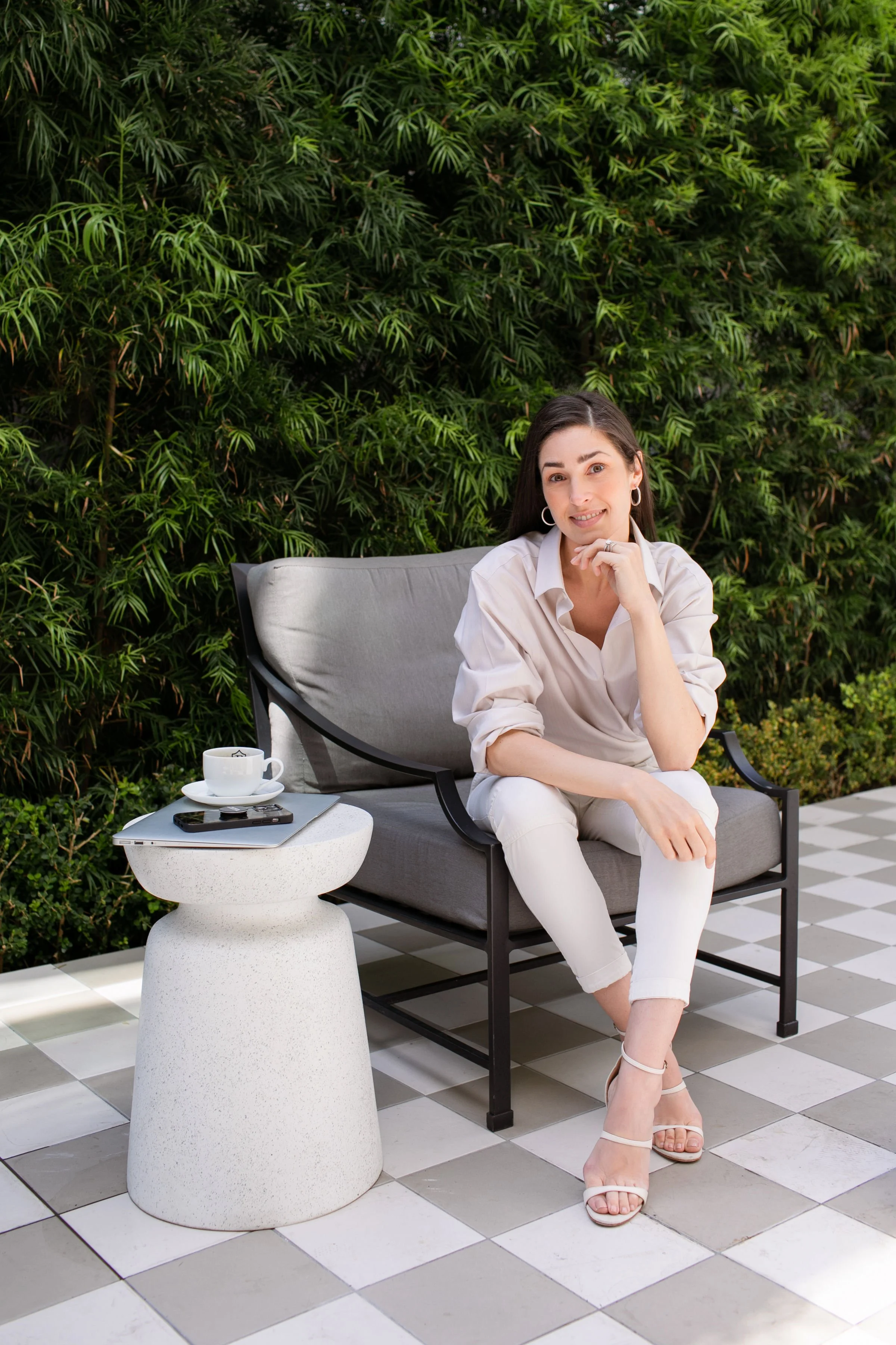 A woman sitting on a gray outdoor sofa with a black metal frame, on a checkered black and white tile patio, in front of a green hedge. She is wearing a light-colored blouse, white pants, and beige high heels, and has a slight smile. On a small textured white side table next to her, there is a coffee cup on a saucer, a smartphone, and a closed laptop.