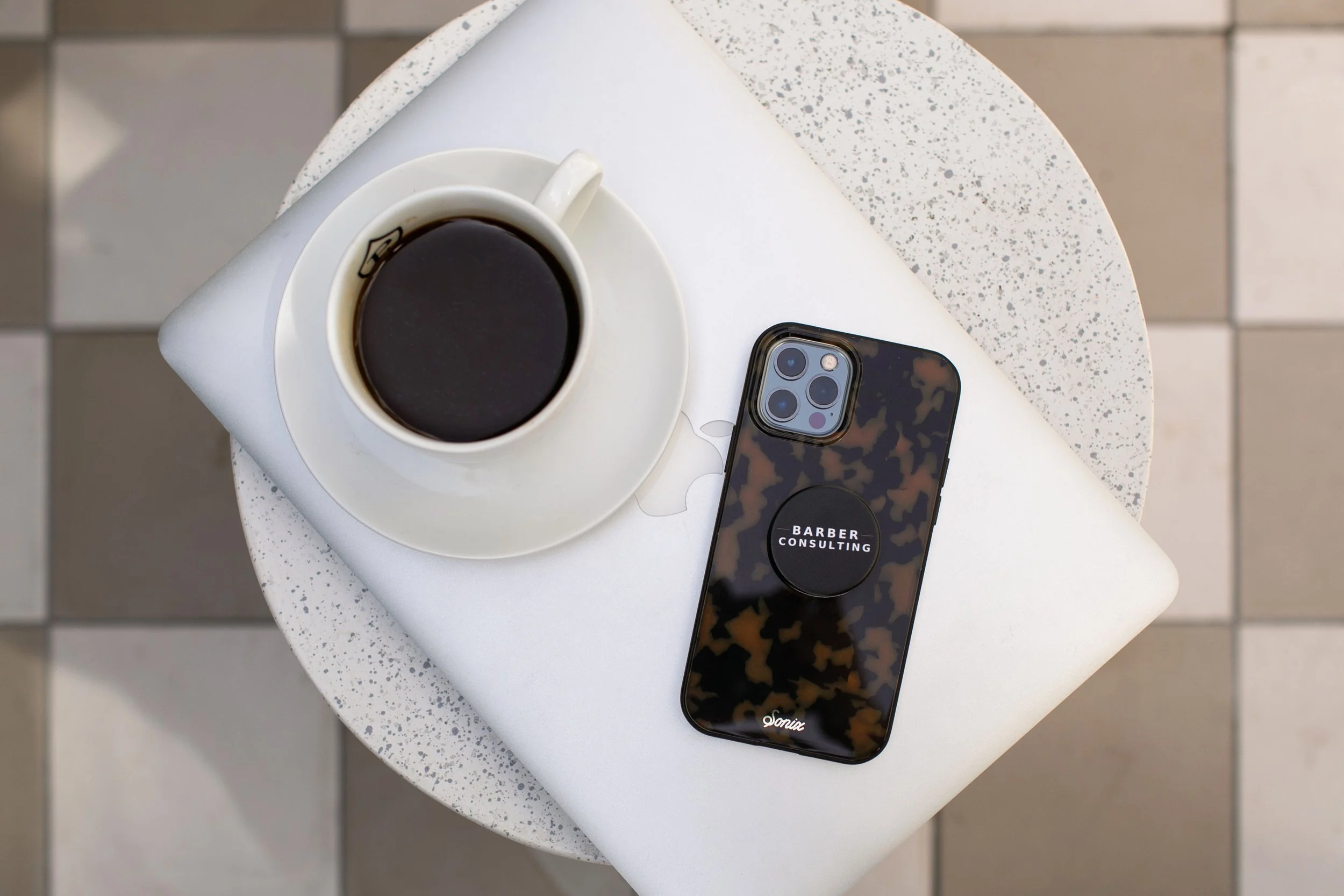 A top-down view of a table with a notebook, a smartphone with a pop socket that says "Barber Consulting" and a coat of arms pattern, a white coffee cup filled with black coffee, all arranged on a terrazzo table in a cafe setting.
