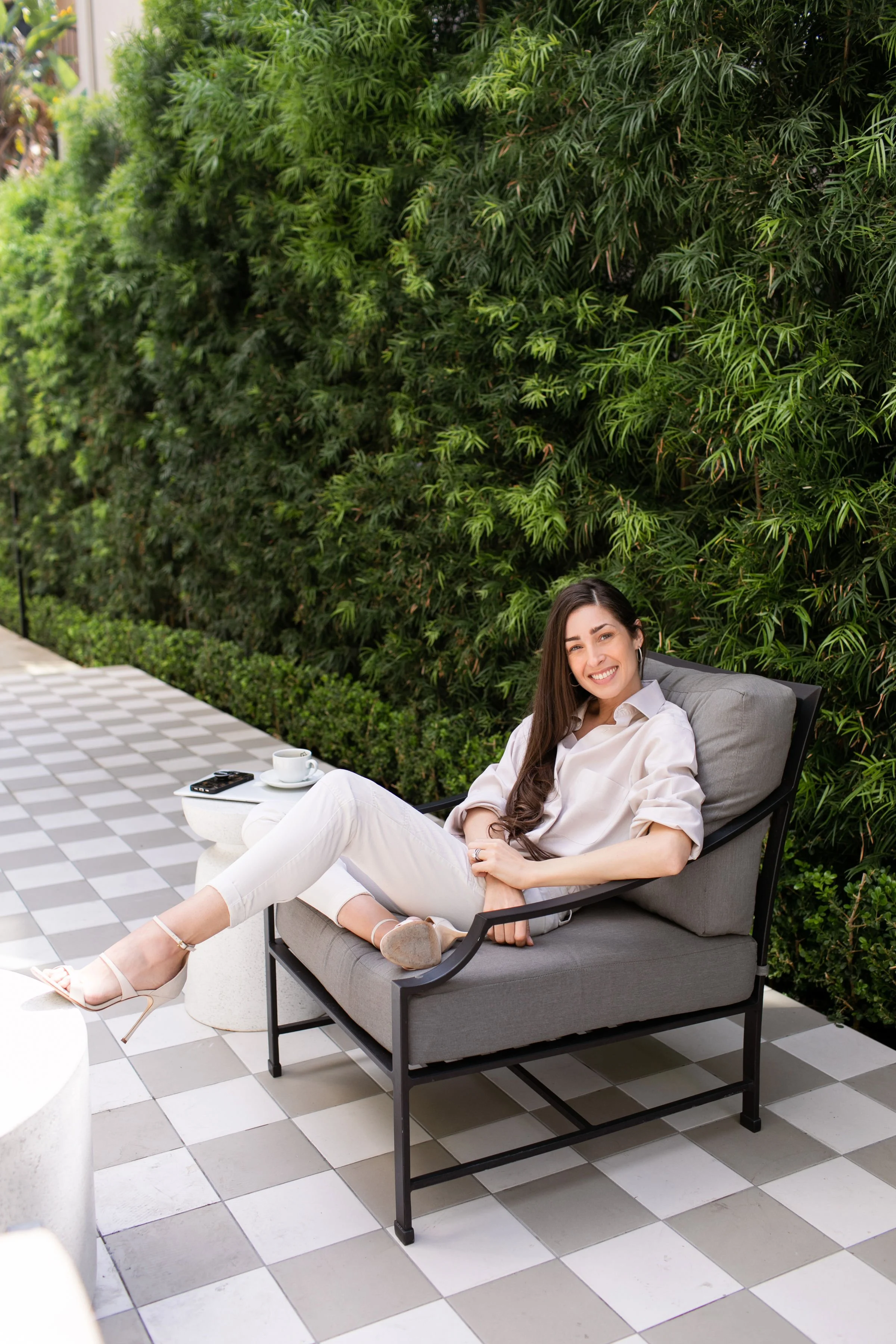 A woman is sitting on a gray outdoor sofa with gray cushions, smiling and sitting relaxed. She is dressed in an off-white shirt and white pants, with beige high heels. Next to her, there is a small white side table holding a white coffee cup, a saucer, and a remote control. The background features a tall green hedge and a checkered tile floor.