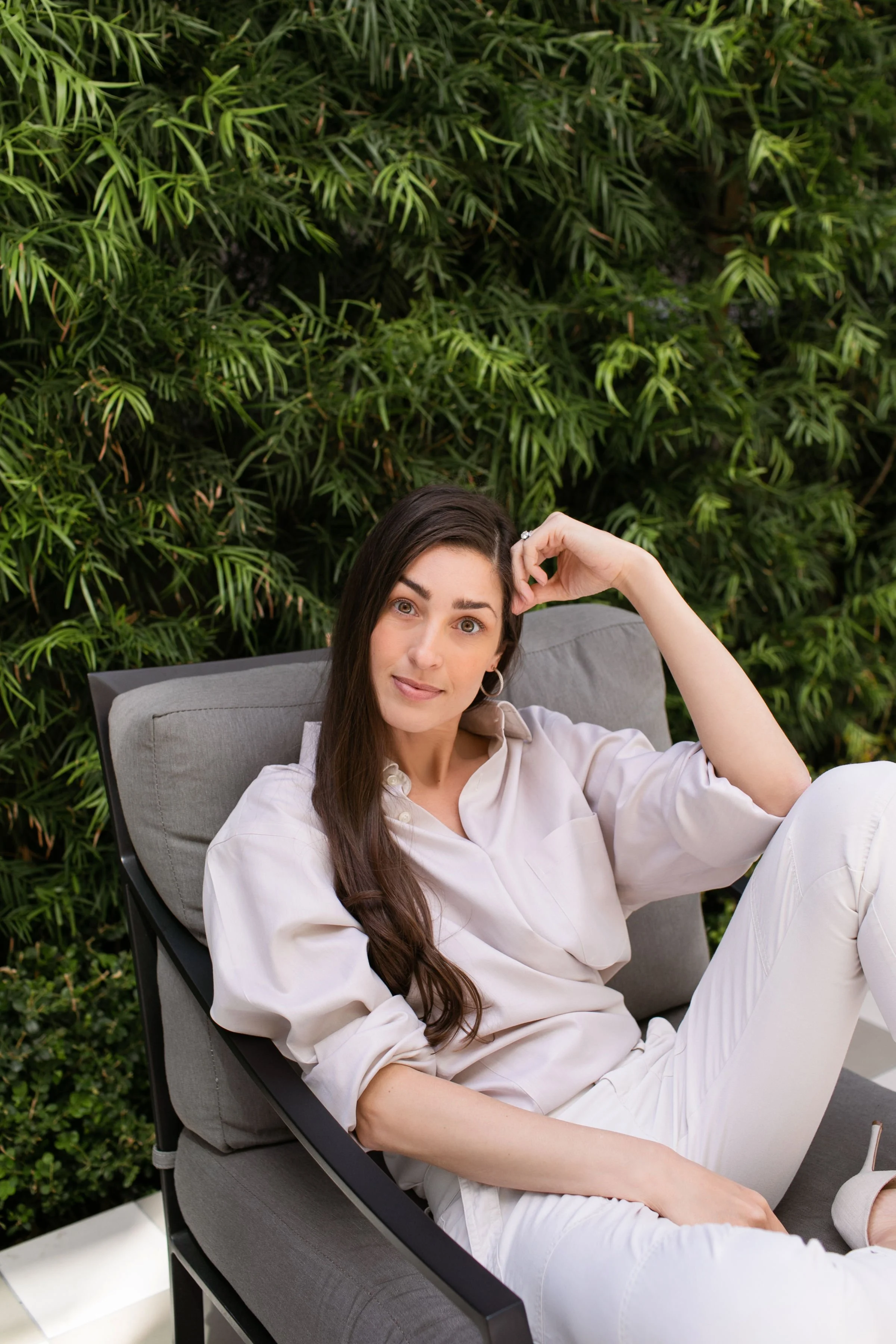 A woman with long dark hair, wearing a cream-colored shirt, sitting on a gray cushioned outdoor lounge chair, surrounded by green foliage. She is looking at the camera with a relaxed expression.