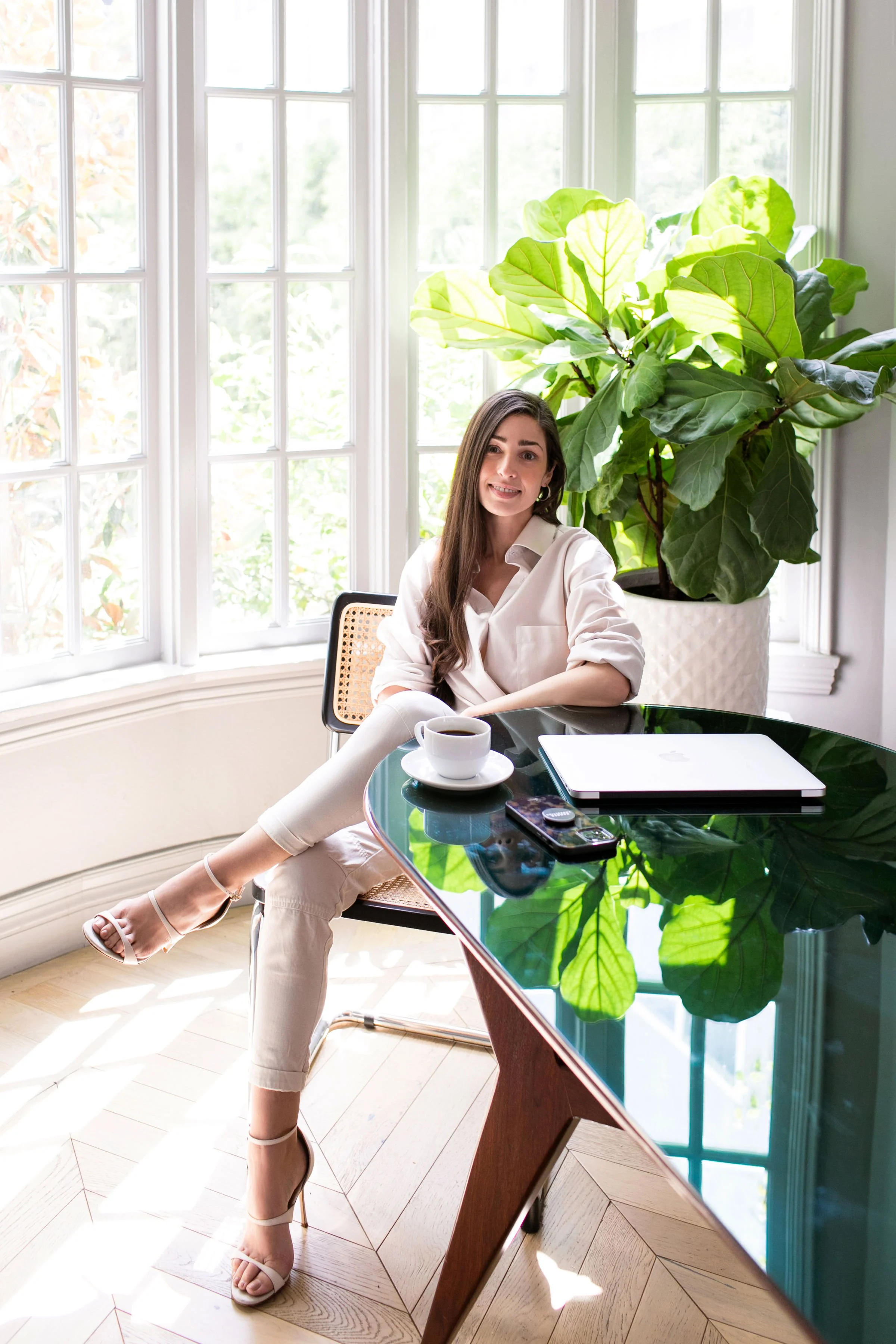 A woman sitting at a glass-top table with a cup of coffee, a closed laptop, and a remote control in front of her. There is a large green plant behind her and large windows letting in natural light.