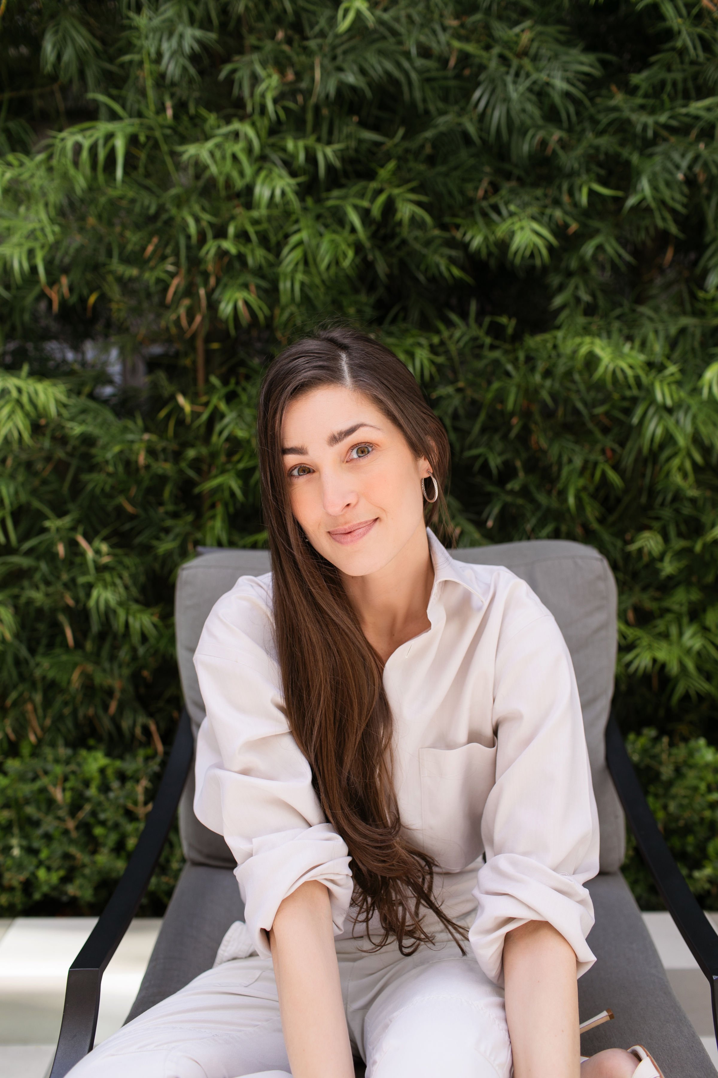 A woman with long brown hair, wearing a white shirt, sitting on a lounge chair outdoors with green foliage in the background.
