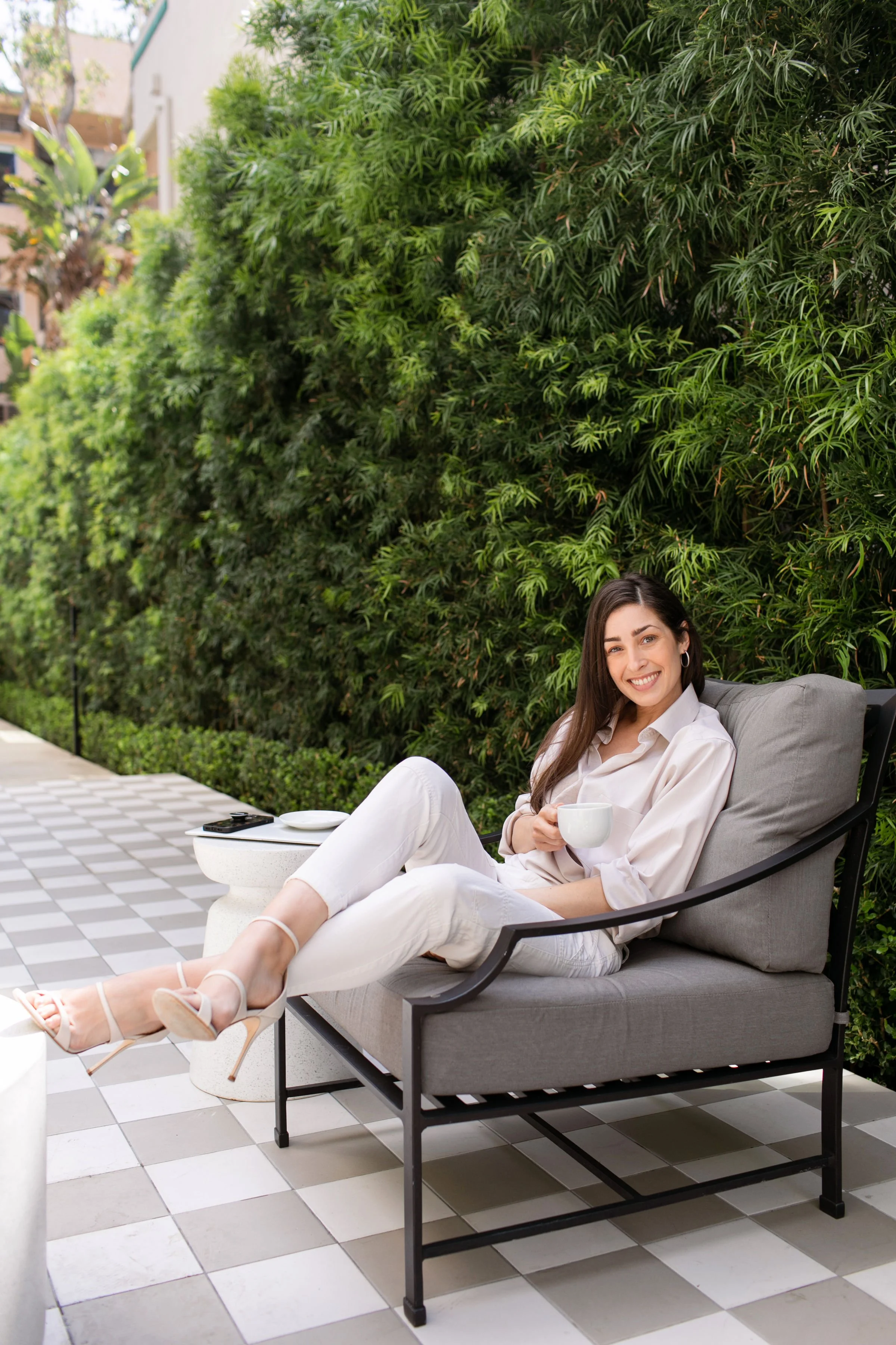 A woman sitting on a patio couch, holding a mug, smiling, with lush green bushes behind her.