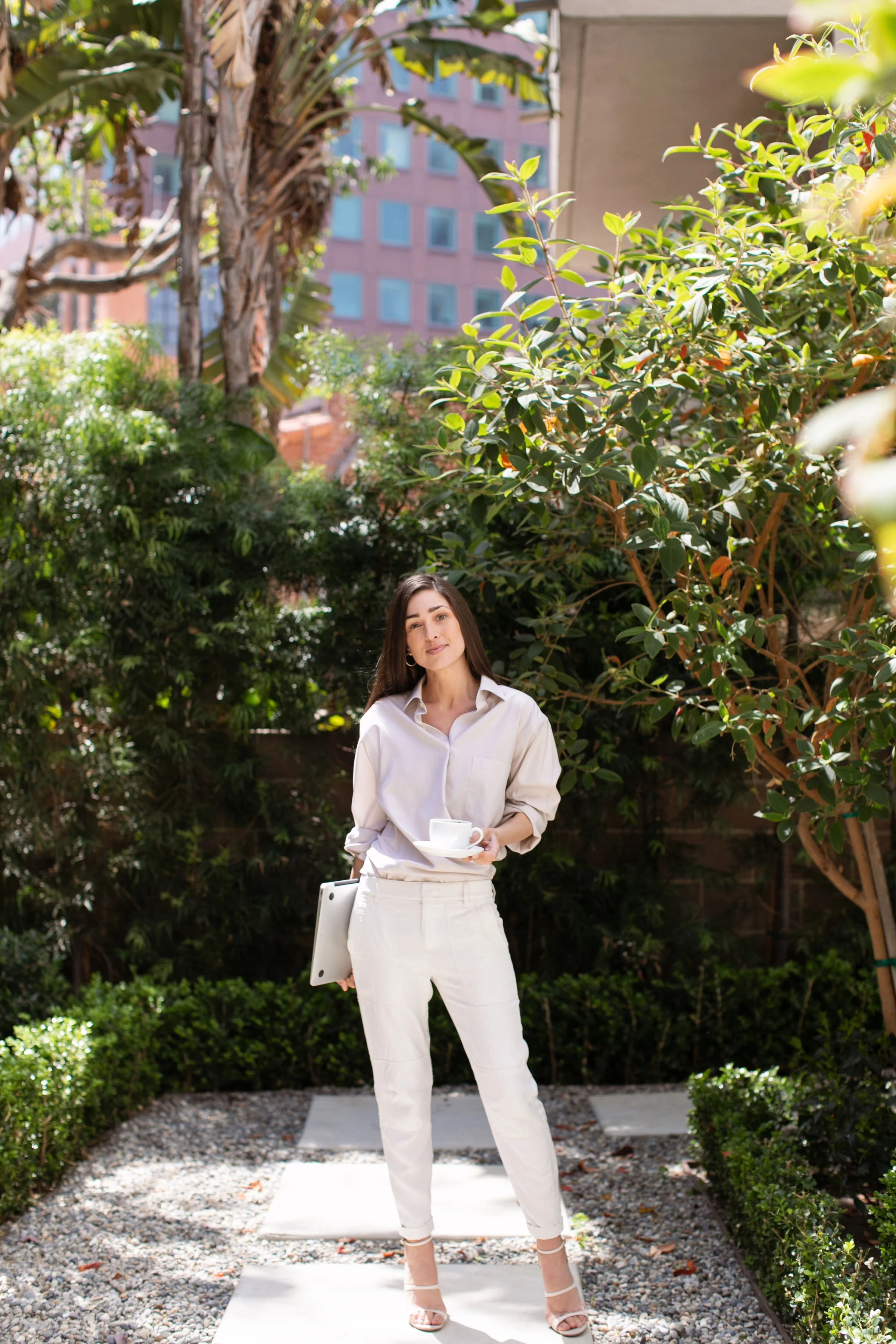 A woman standing outdoors in a garden with lush green foliage, holding a white coffee cup and saucer, carrying a tablet under her arm, dressed in an off-white shirt and pants, with a city building visible in the background.