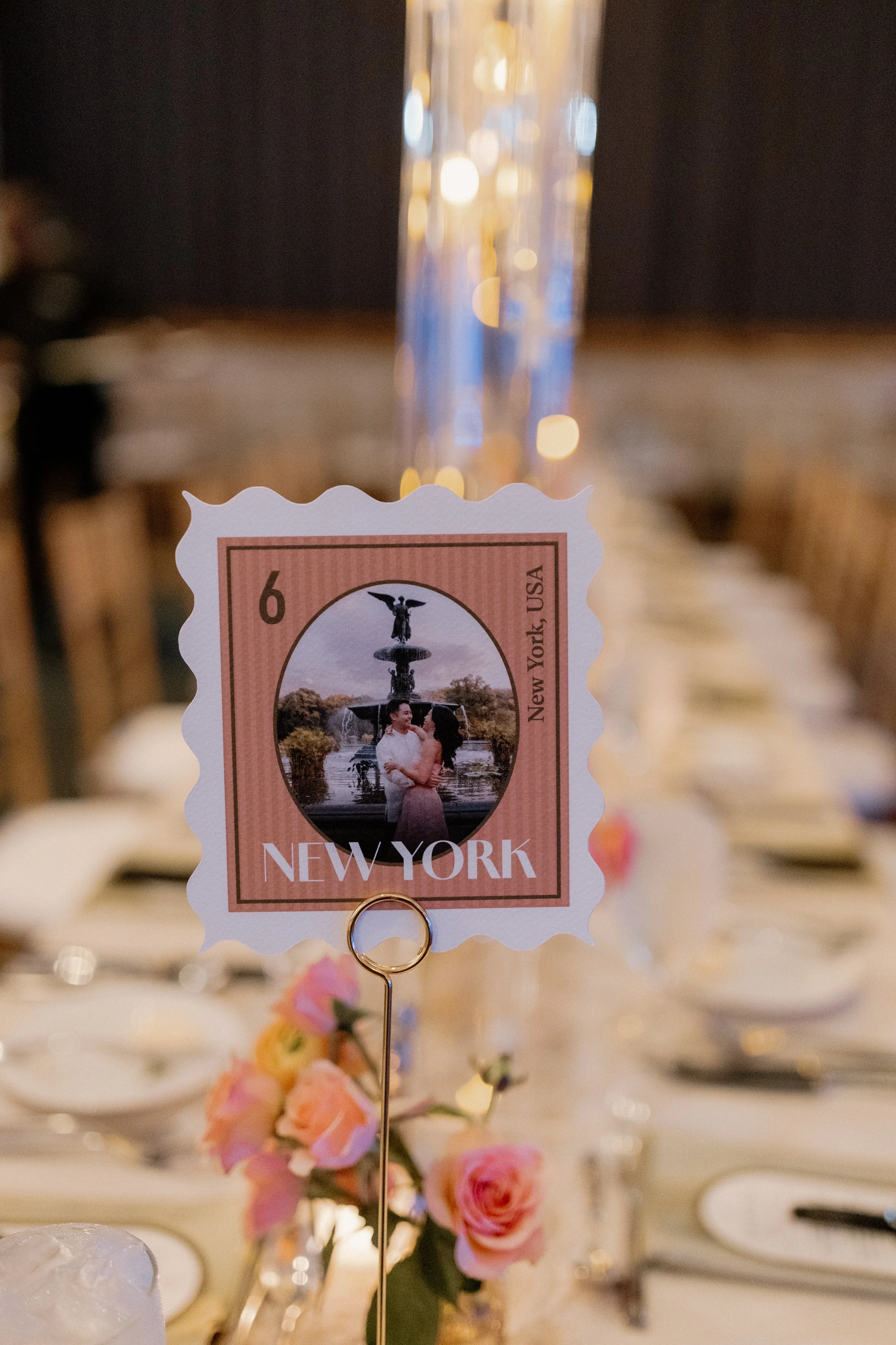 A close-up photo of a New York-themed wedding table centerpiece featuring a pink postage stamp depiction of a fountain with two women, placed above pink roses and surrounded by dining table settings in a dimly lit venue.