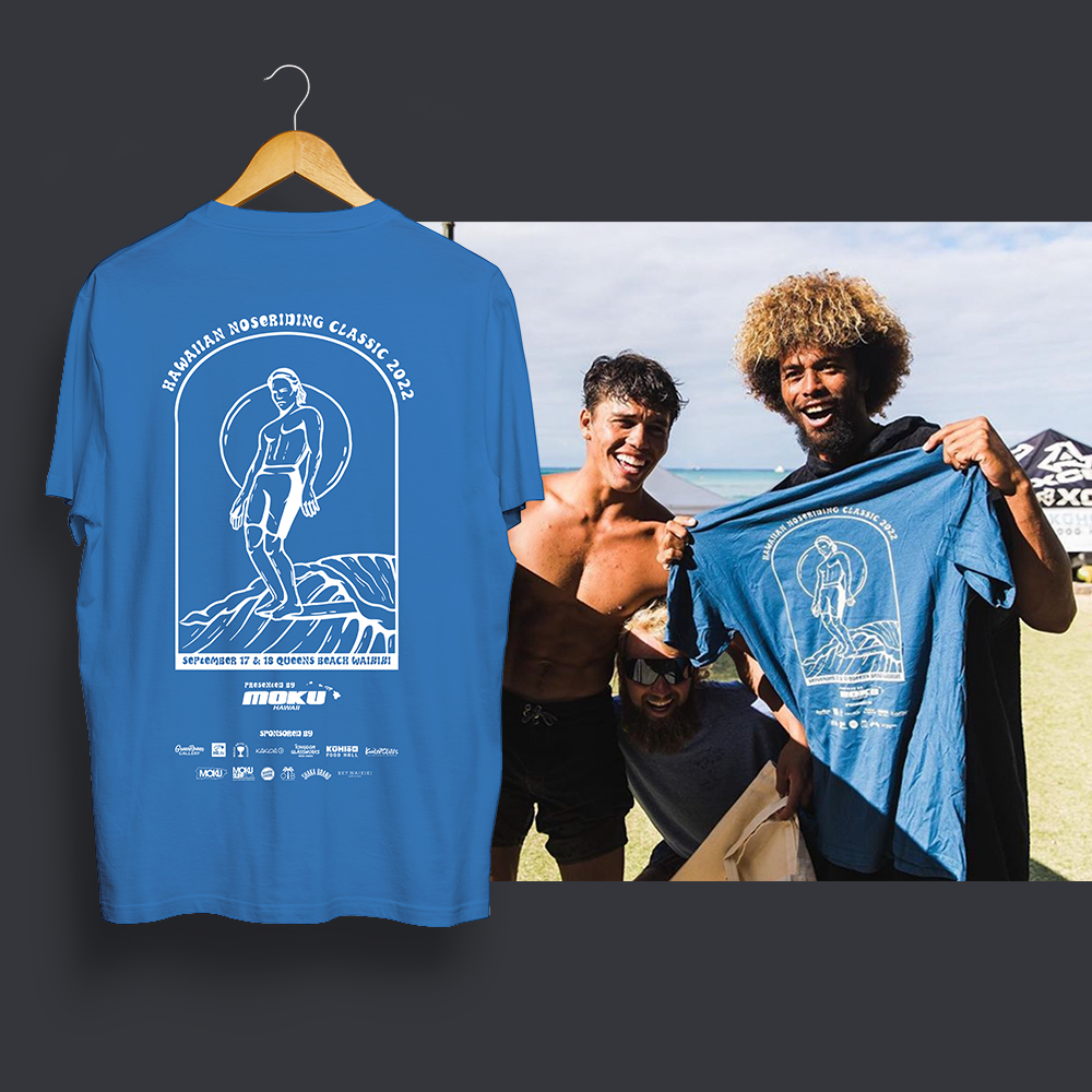A group of three men at a beach event showing off a blue event shirt with a surfer design and text, with one man holding the shirt and another smiling at the camera.