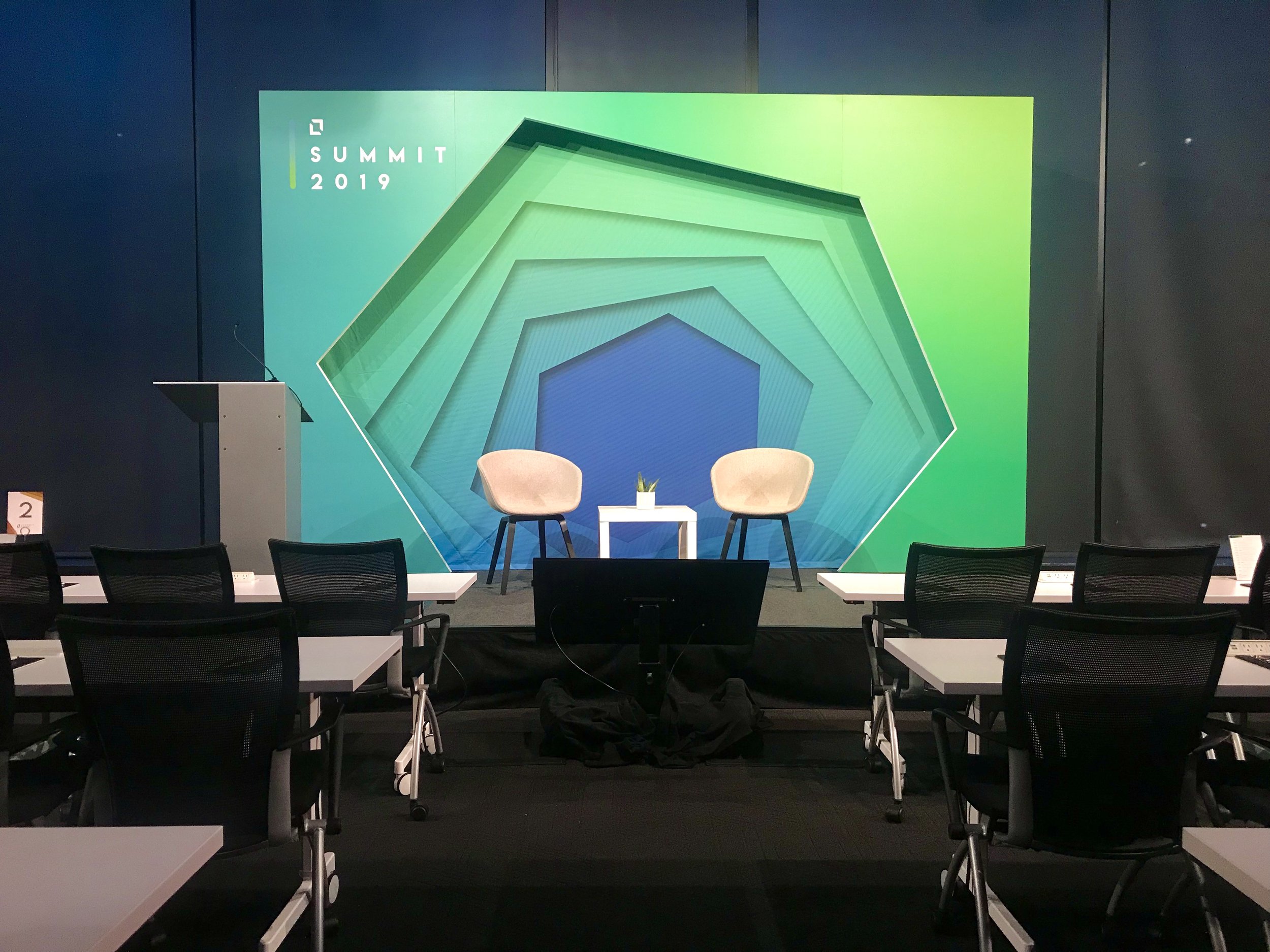 Empty conference stage with two beige chairs, a small white table with a plant, and a large green and blue geometric backdrop at a Summit 2019 event.