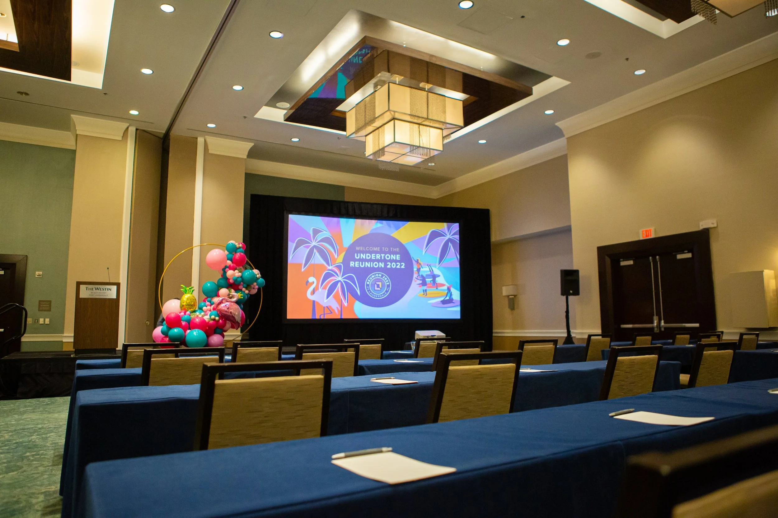 Conference room at The Westin hotel set up for Undertone Reunion 2022, with rows of chairs and tables, a stage with a colorful screen, balloons with tropical designs, and ceiling lights.