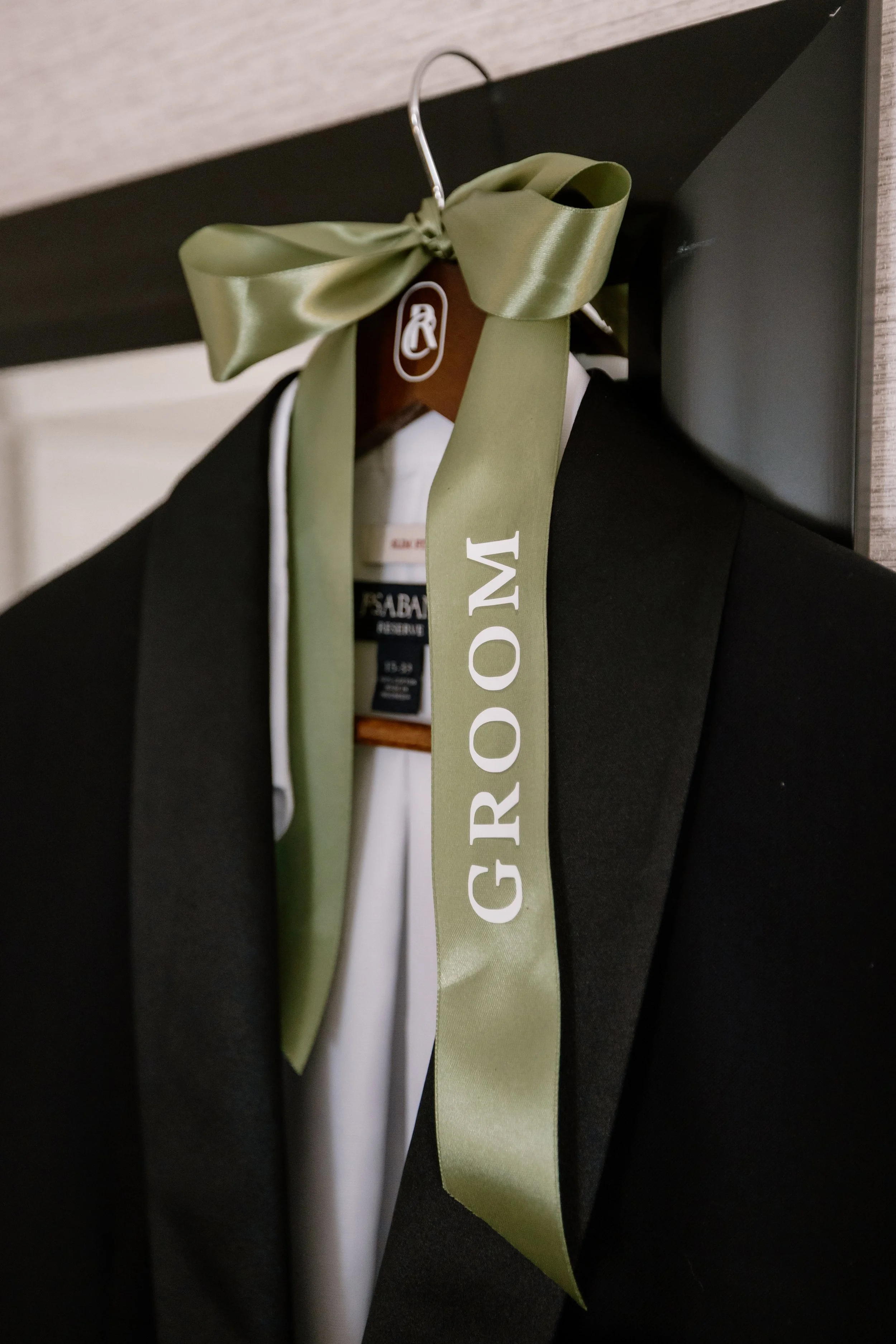 A tuxedo jacket hanging on a hanger decorated with a green ribbon that has the word 'GROOM' printed on it.