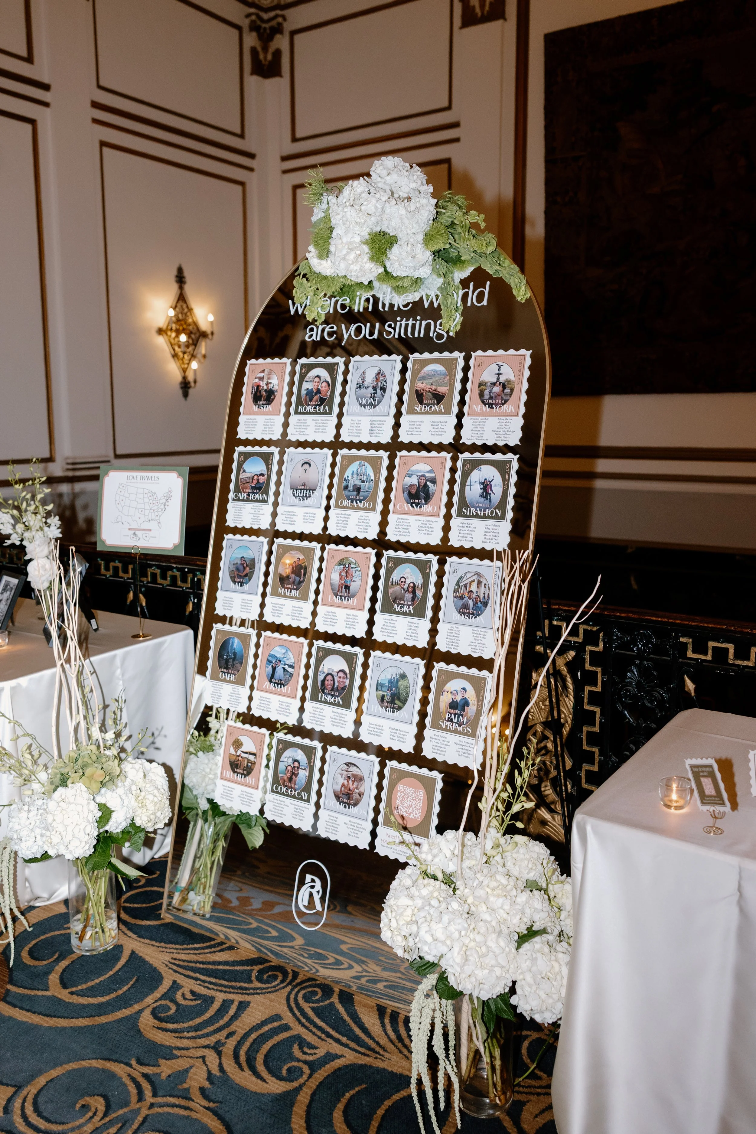 Wedding seating chart display with photos and names of guests, decorated with white hydrangeas and greenery, in a decorated ballroom.