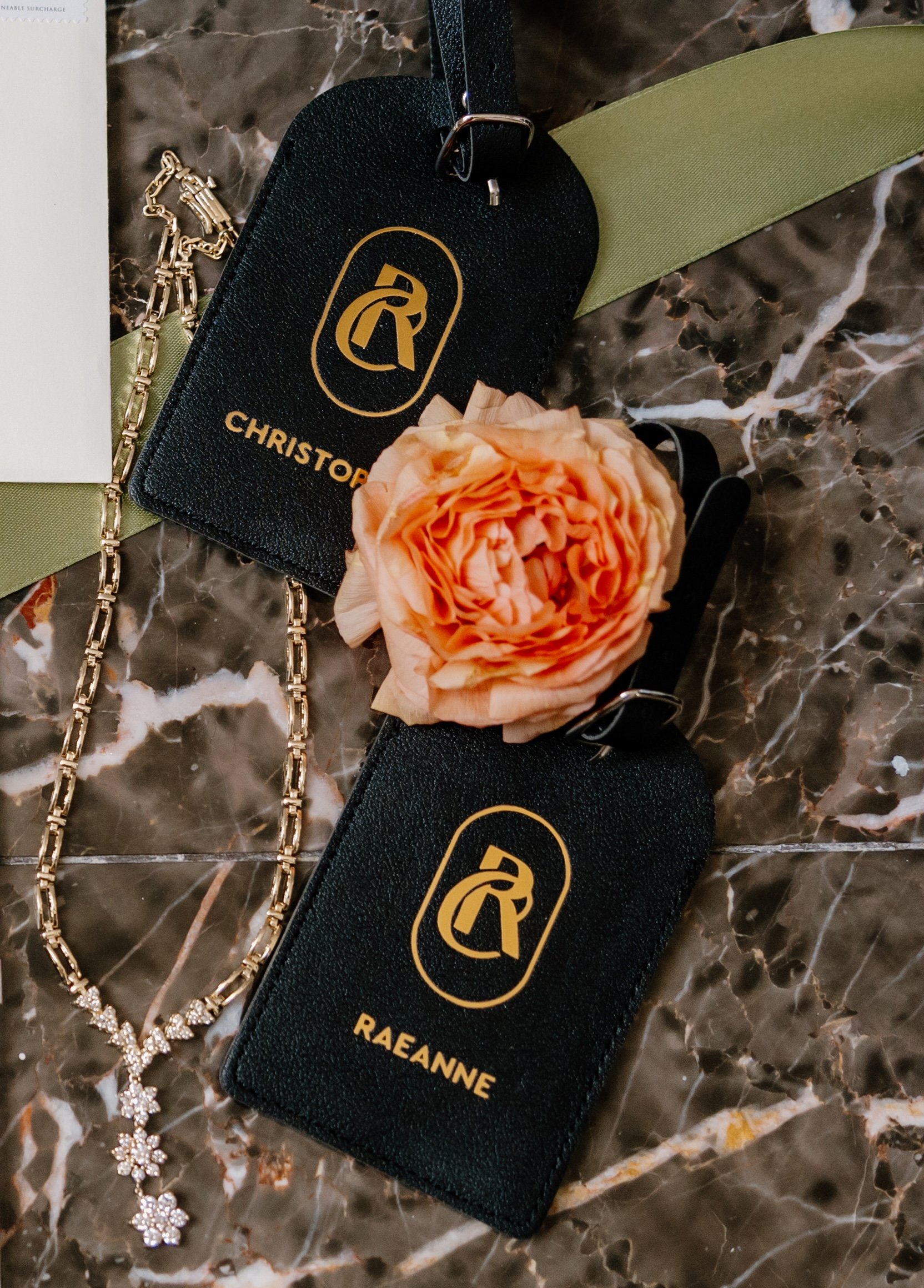 Black tags with gold lettering, one reads 'CHRISTOPHER' and the other 'RAEANN', with a gold logo, positioned on a marble surface with a large peach-colored flower, a gold chain necklace, and a green ribbon.