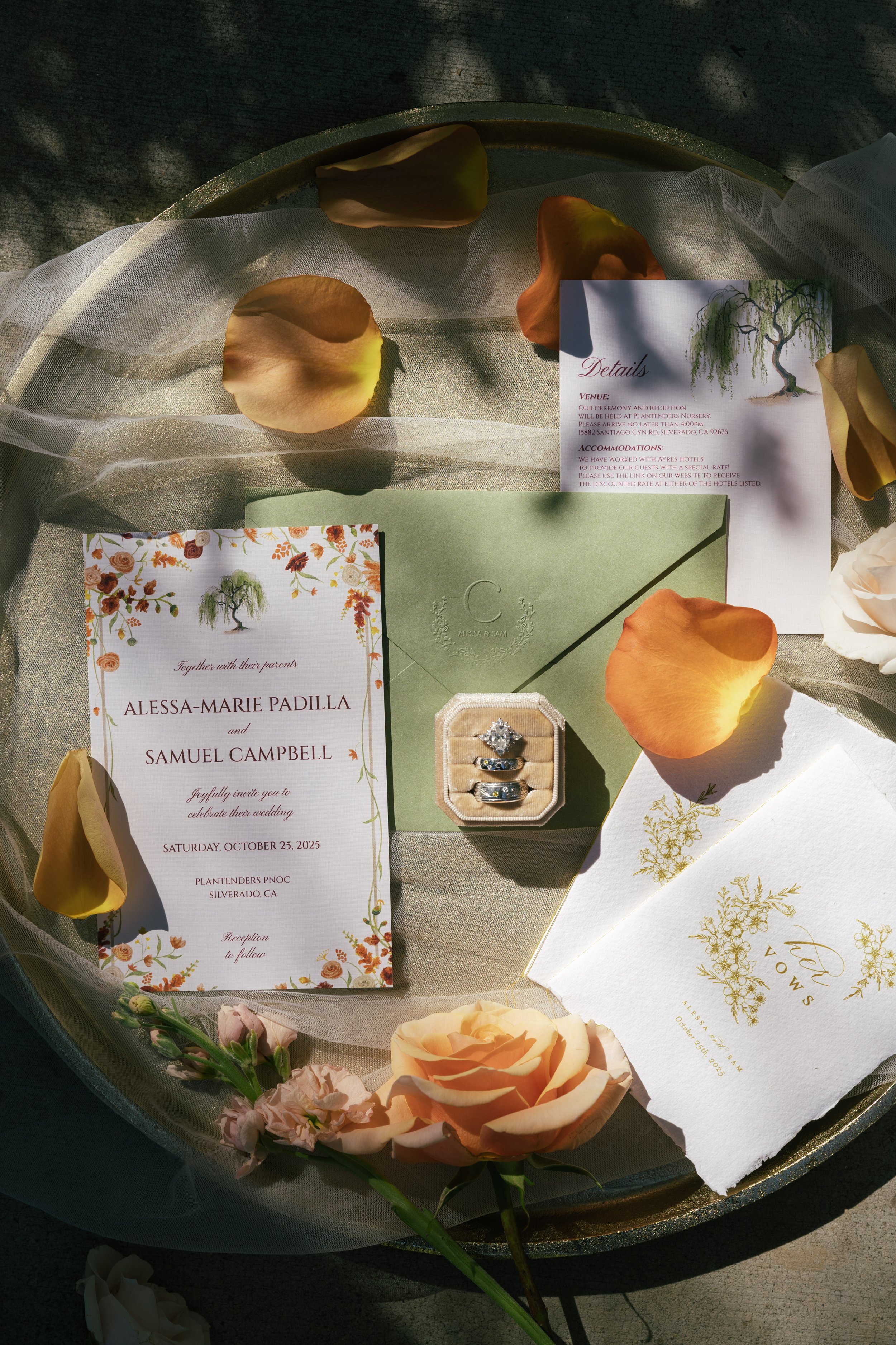 A wedding invitation display with rings, floral elements, and informational cards in a tray, decorated with rose petals and flowers.