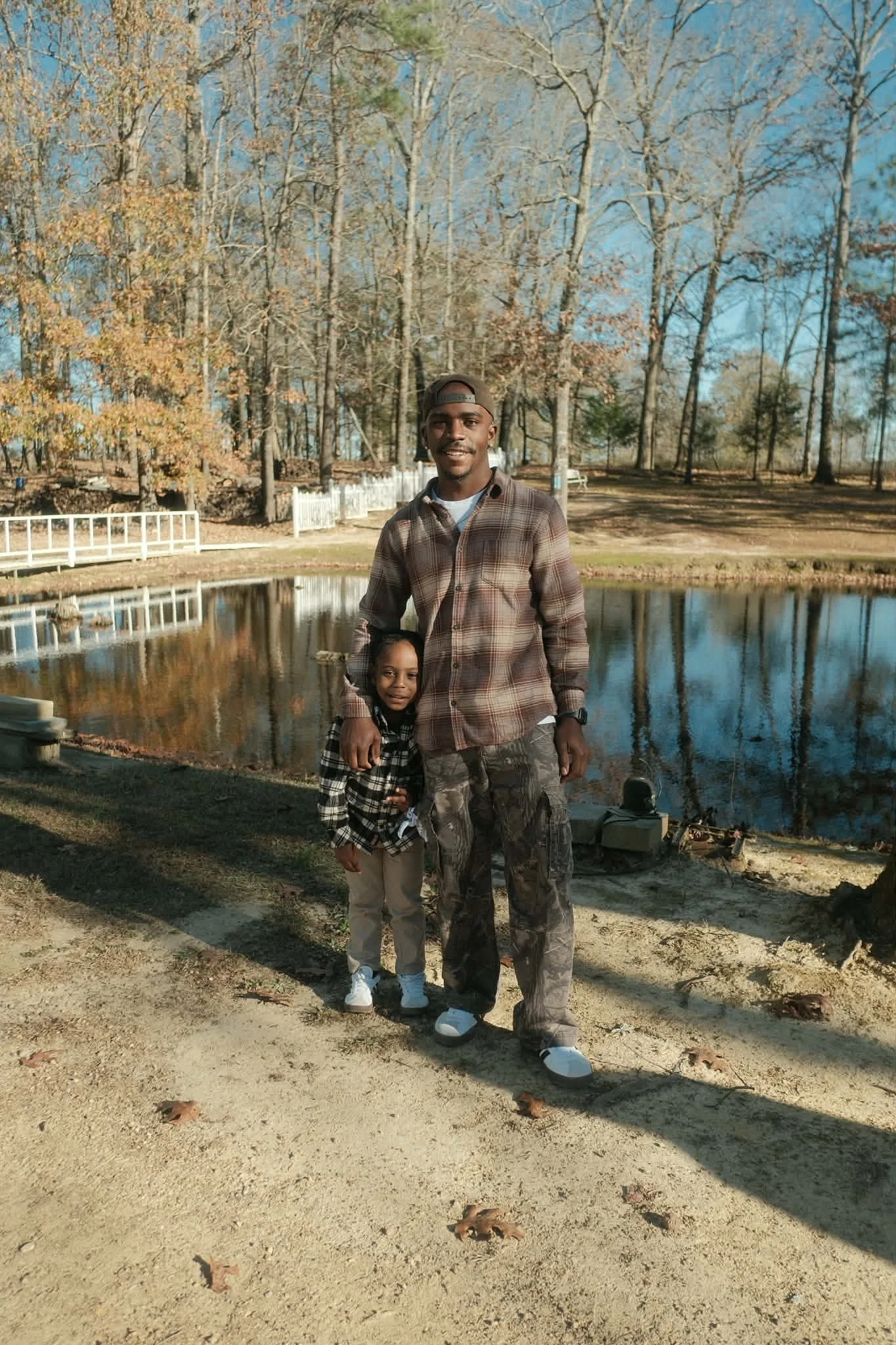 A man and a young girl standing by a pond in a park during autumn. The man has his arm around the girl, and both are smiling at the camera. The background features leafless trees and a white fence.