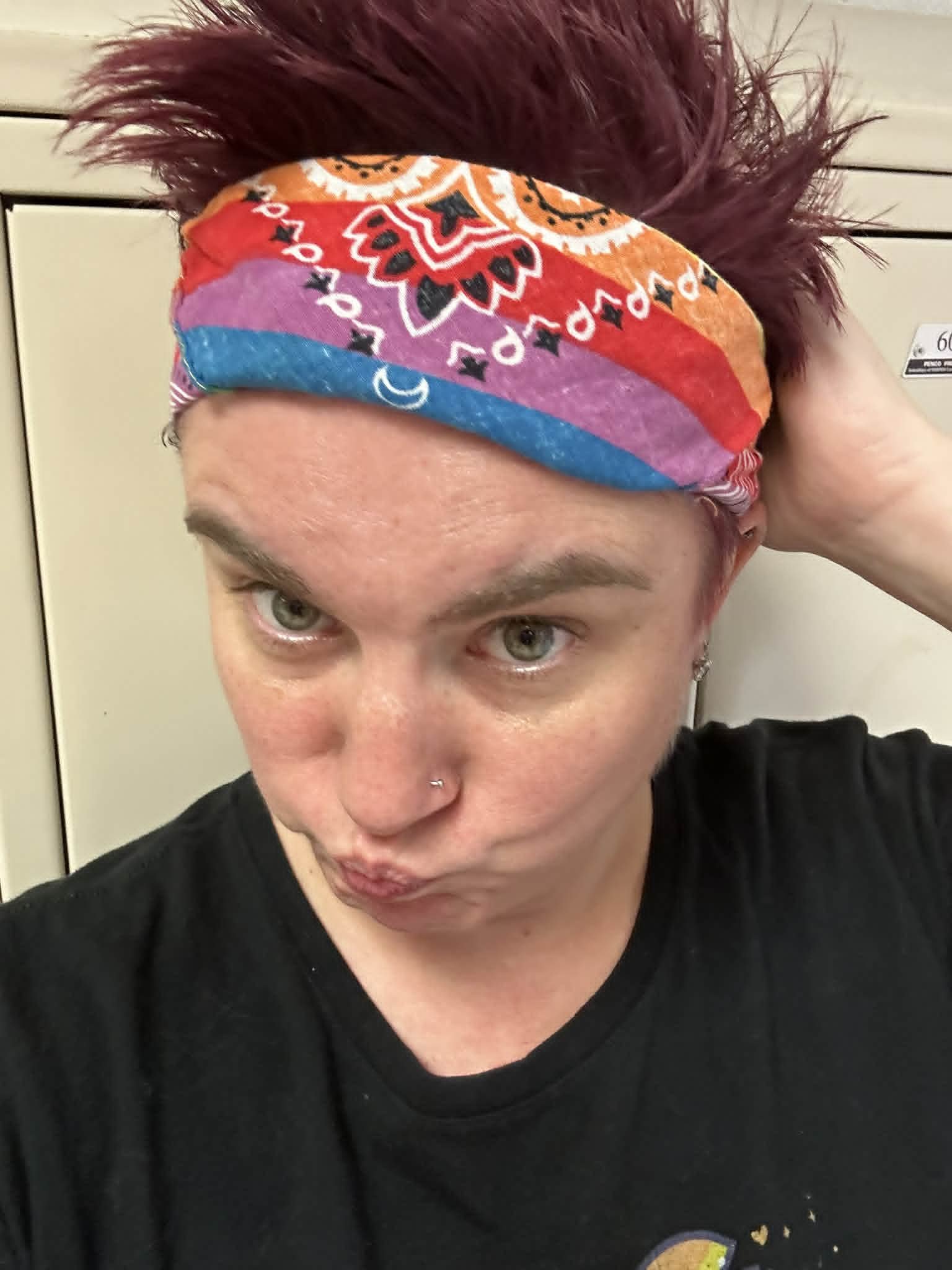A person with short red hair wearing a colorful bandana and a black T-shirt, posing with a playful facial expression. The background features a beige-colored locker.