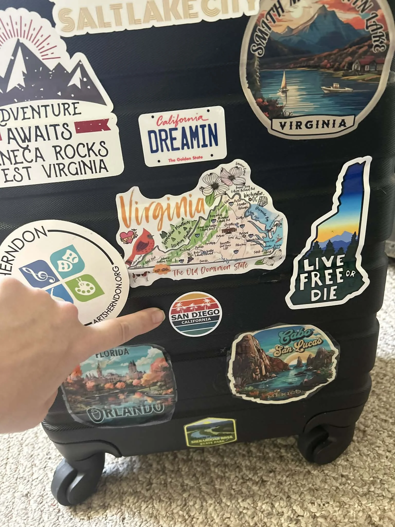 Luggage decorated with travel stickers including Virginia map, San Diego logo, Orlando, Florida, Cabo San Lucas, and a sticker reading "Live Free or Die".