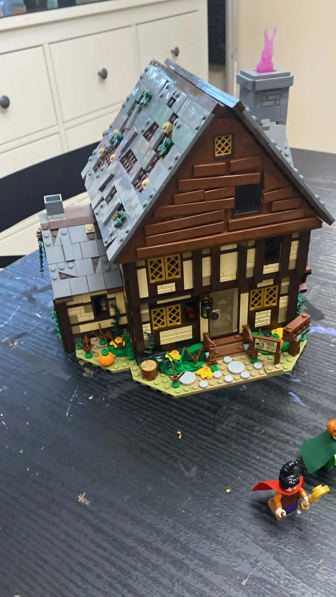 Lego model house with brown walls, gray shingled roof, and front porch, placed on a dark table with Lego figures nearby.