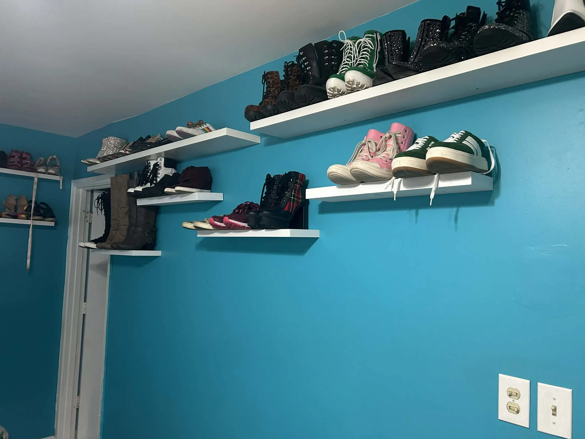 Wall with white shelves holding various pairs of shoes and boots in a room with bright blue walls.