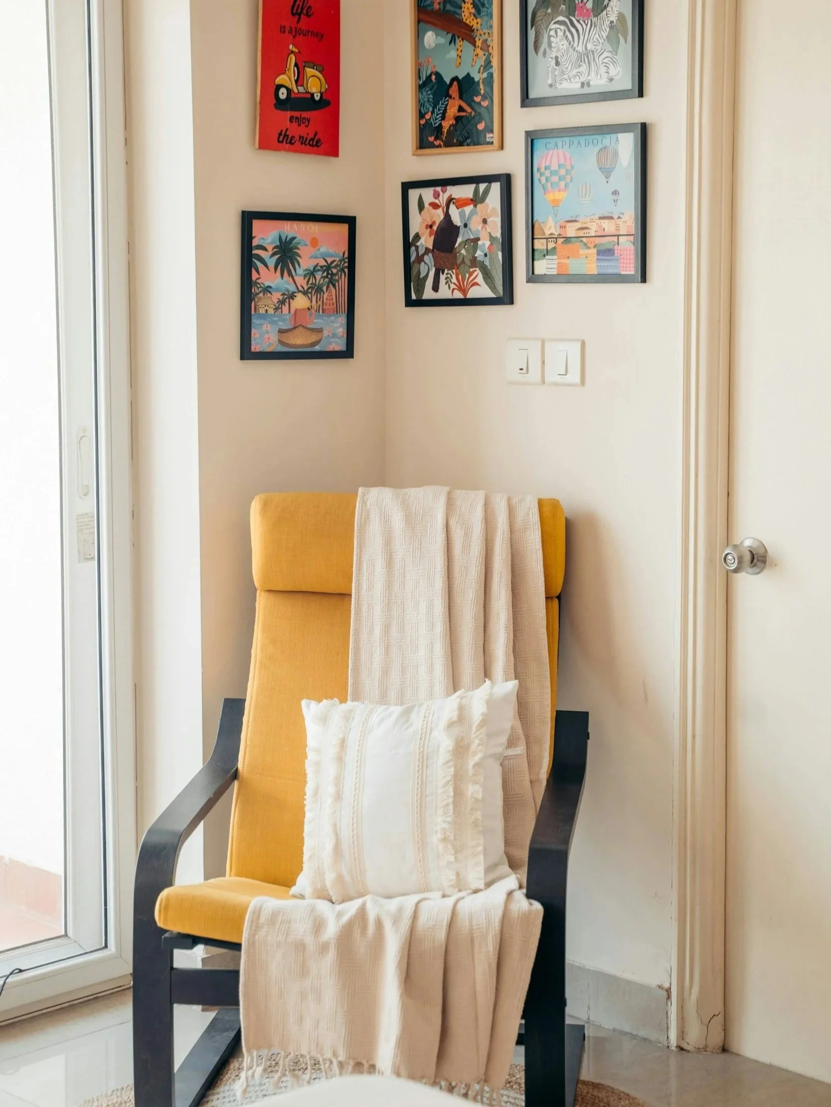 Yellow armchair with beige blankets and pillow, positioned next to a sliding glass door in a room decorated with framed colorful vacation-themed artwork on the wall.