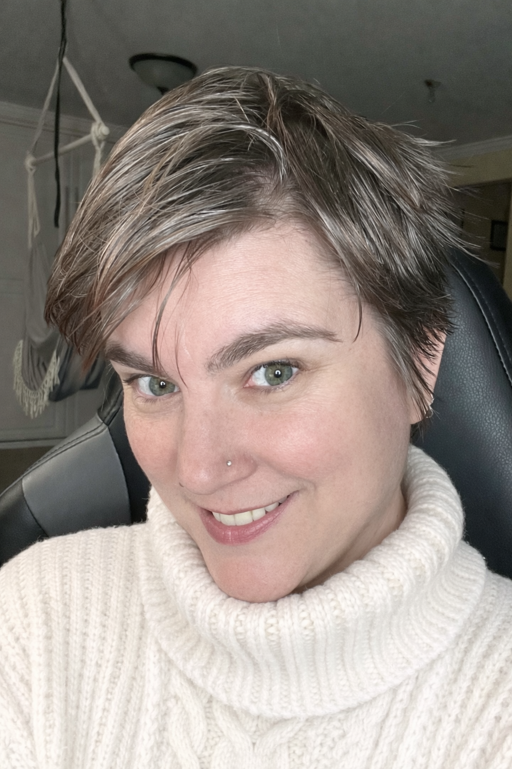 A woman with short, light brown hair and a nose piercing, wearing a cream-colored turtleneck sweater, smiling at the camera, sitting in a black chair inside a room.