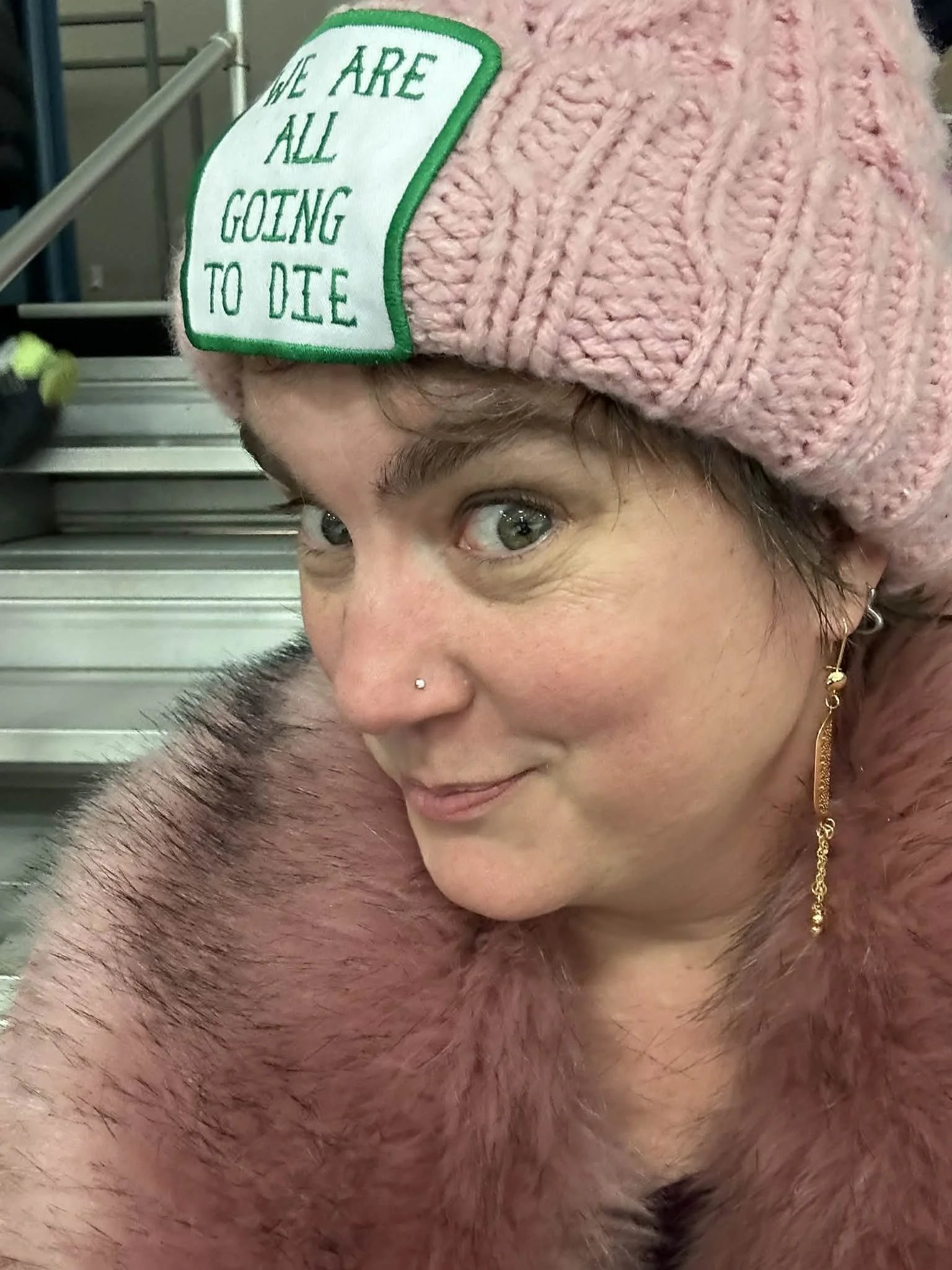 A woman with a nose piercing and gold earring wears a pink knit hat with a patch that says 'We are all going to die.' She is also wearing a pink fur coat and is sitting on metal stairs, smiling slightly.