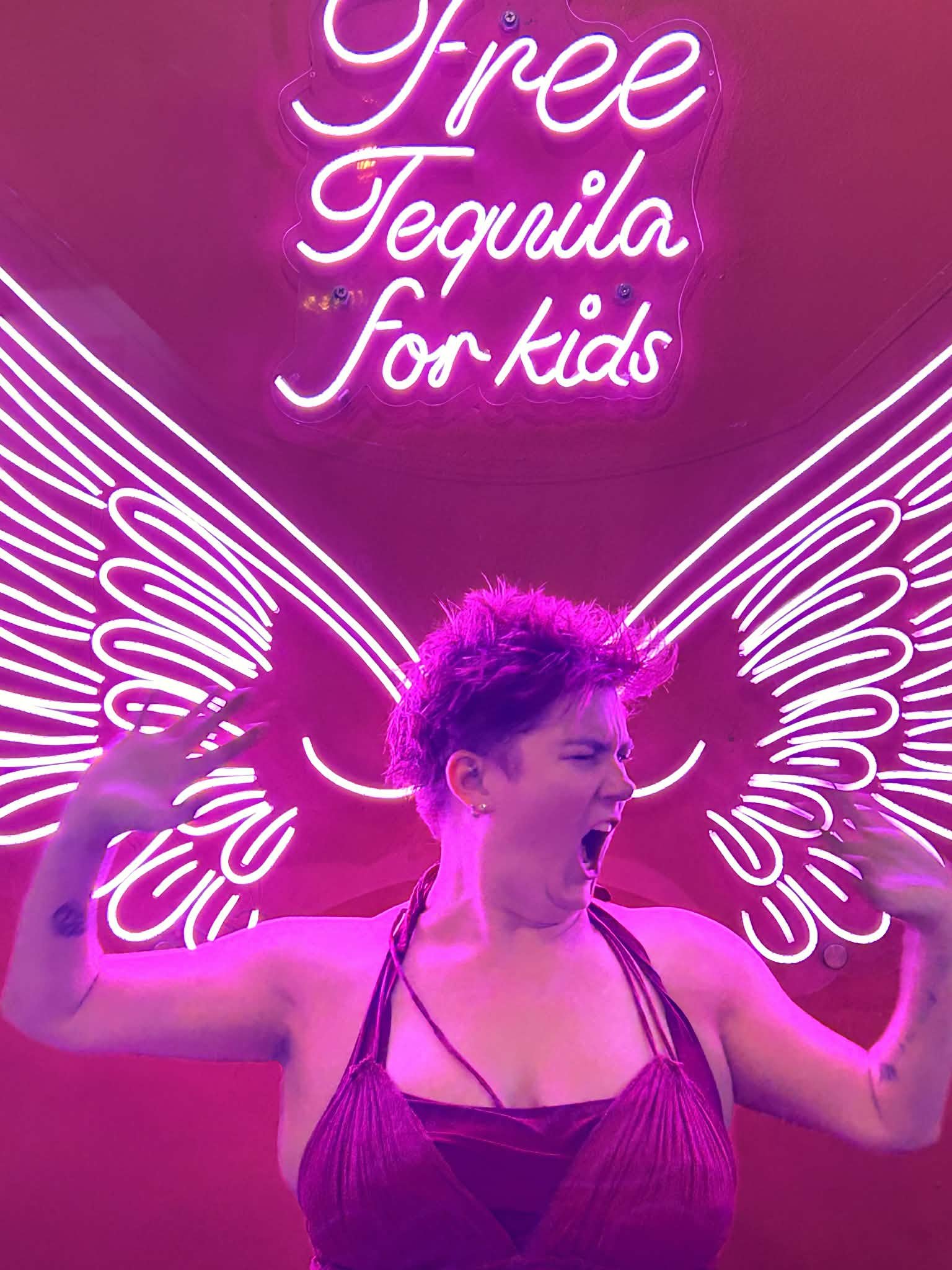 Person making a fierce face with muscular arms raised, pink neon sign above that reads "Free Tequila for Kids", and neon angel wings in the background.