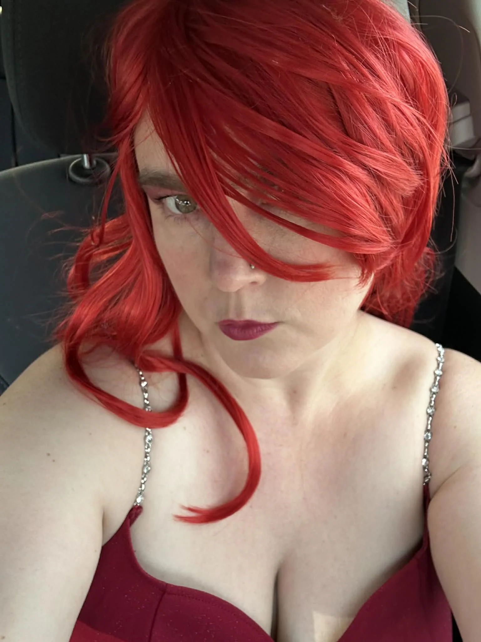 A woman with bright red hair taking a selfie, wearing a red dress with thin, bejeweled straps, seated indoors with dark background.