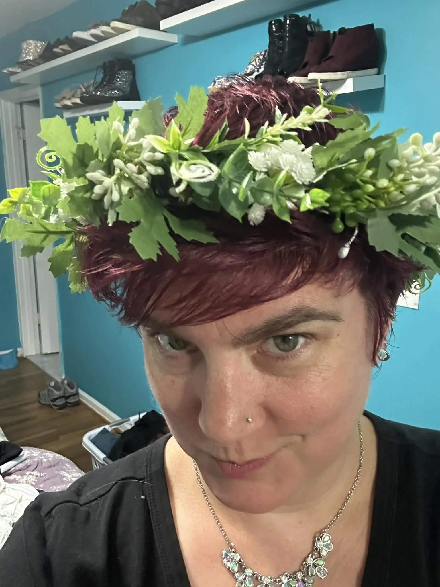 Person with short red hair wearing a floral wreath on her head, a nose piercing, and a necklace, indoors with a teal wall, shoes, and clothes visible in the background.