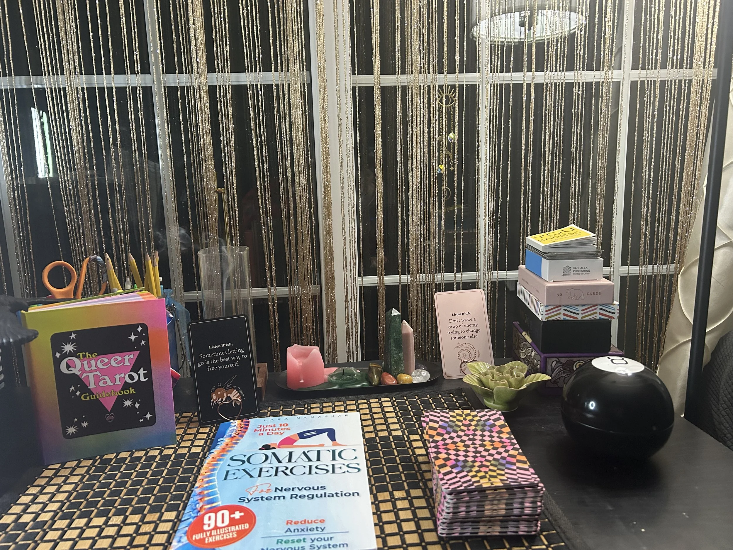 A table with various tarot and oracle cards, small stacks of cards, a book titled 'Somatic Exercises,' decorative crystals, candles, and motivational cards. The background features hanging gold and silver beaded curtains.