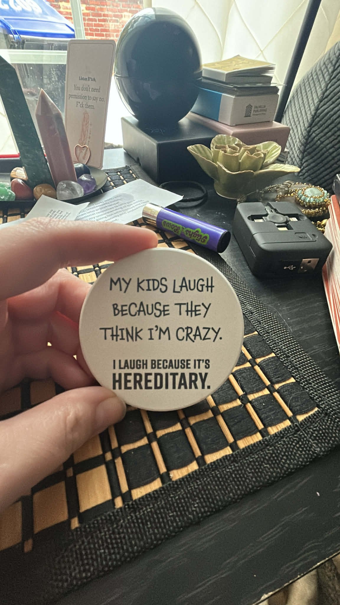 A round white sticker with black text saying, "My kids laugh because they think I’m crazy. I laugh because it’s hereditary." Held between a person's thumb and index finger, with a cluttered desk in the background containing various objects including 