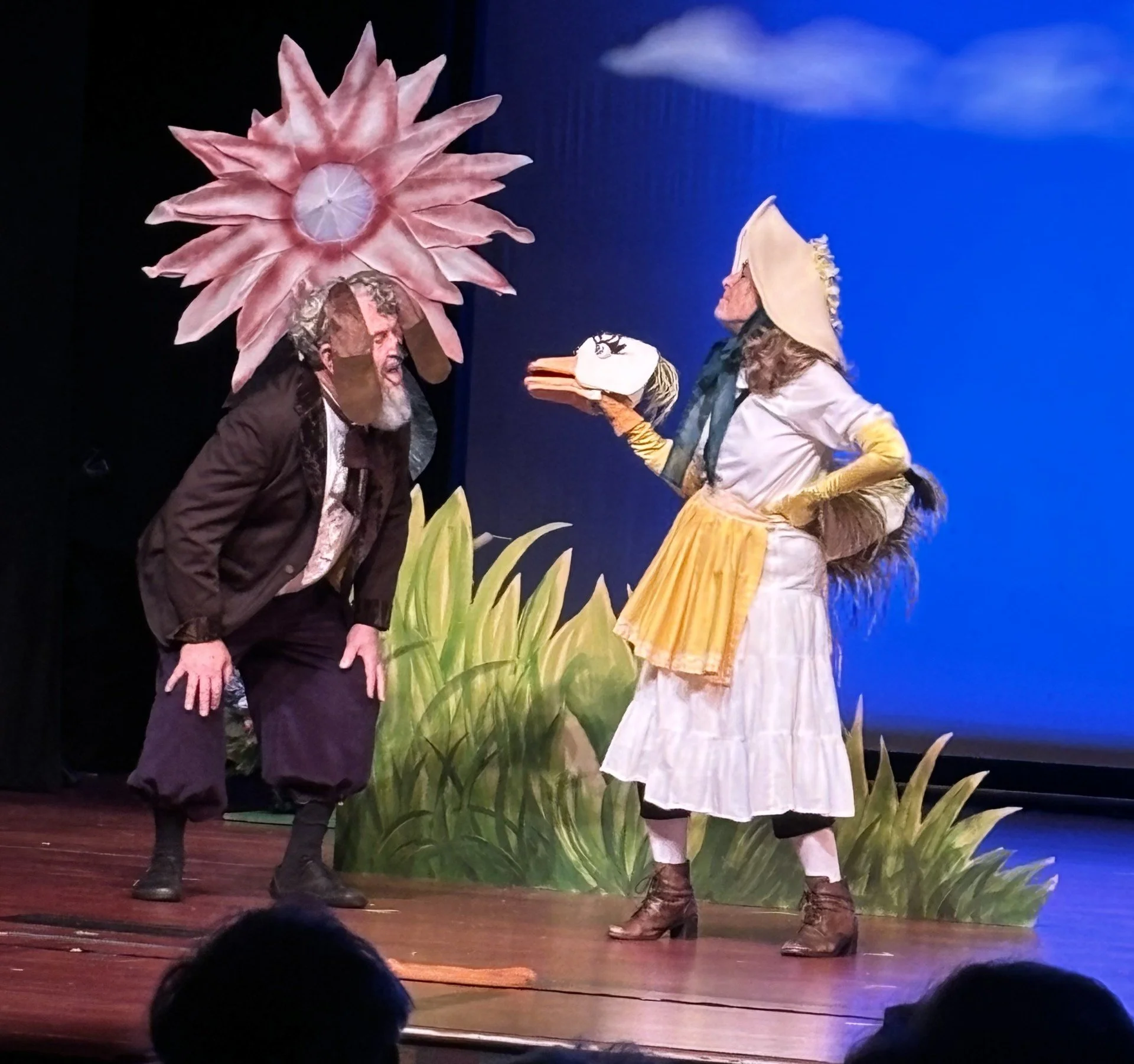 Two actors in costume performing on stage: one dressed as an elderly man with a beard and dark clothing, leaning forward, and the other as a woman wearing a large bonnet, yellow apron, and holding a goose-shaped object, with a backdrop of a large flower and greenery.