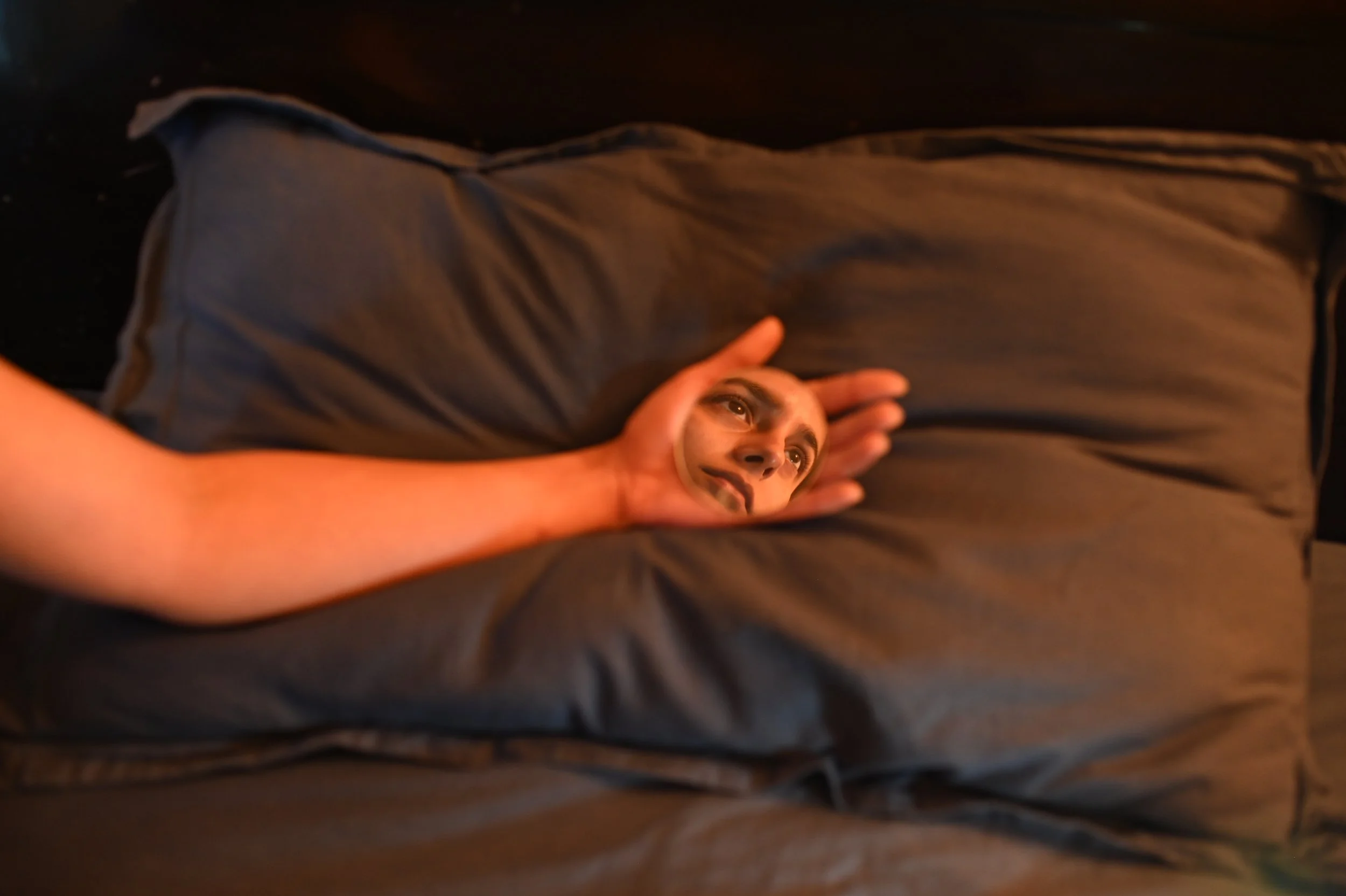 Person lying on a bed holding a small mirror that shows their face, with dark brown bedding.