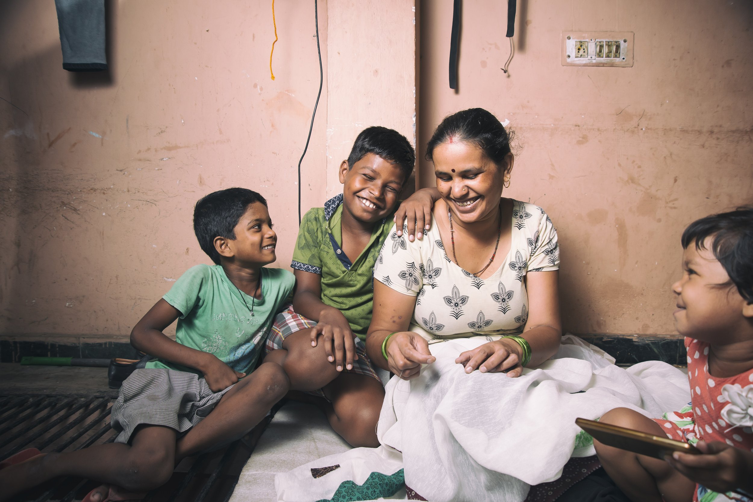 A woman and four children sitting on the floor, smiling and laughing together in a room with pink walls.