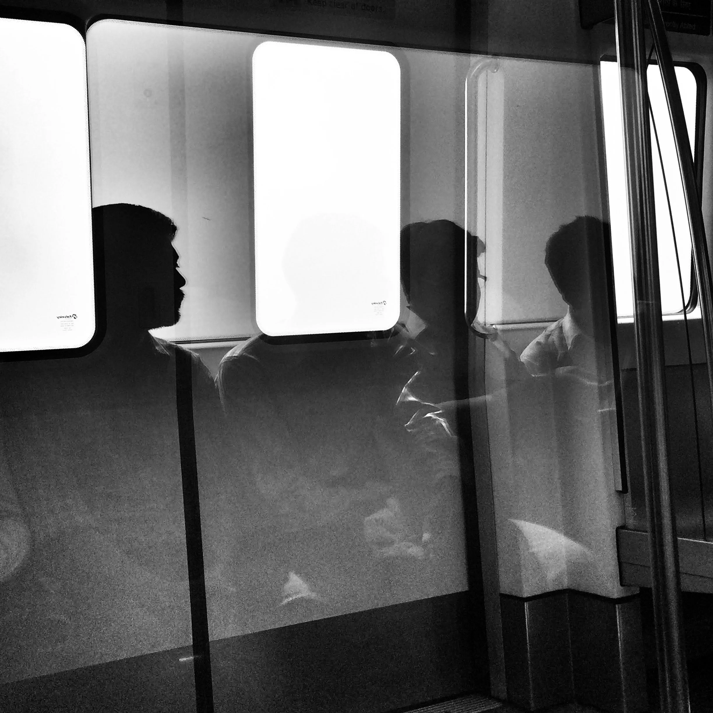 Silhouettes of four people seated on a bus, seen through a window with reflections, in black and white.