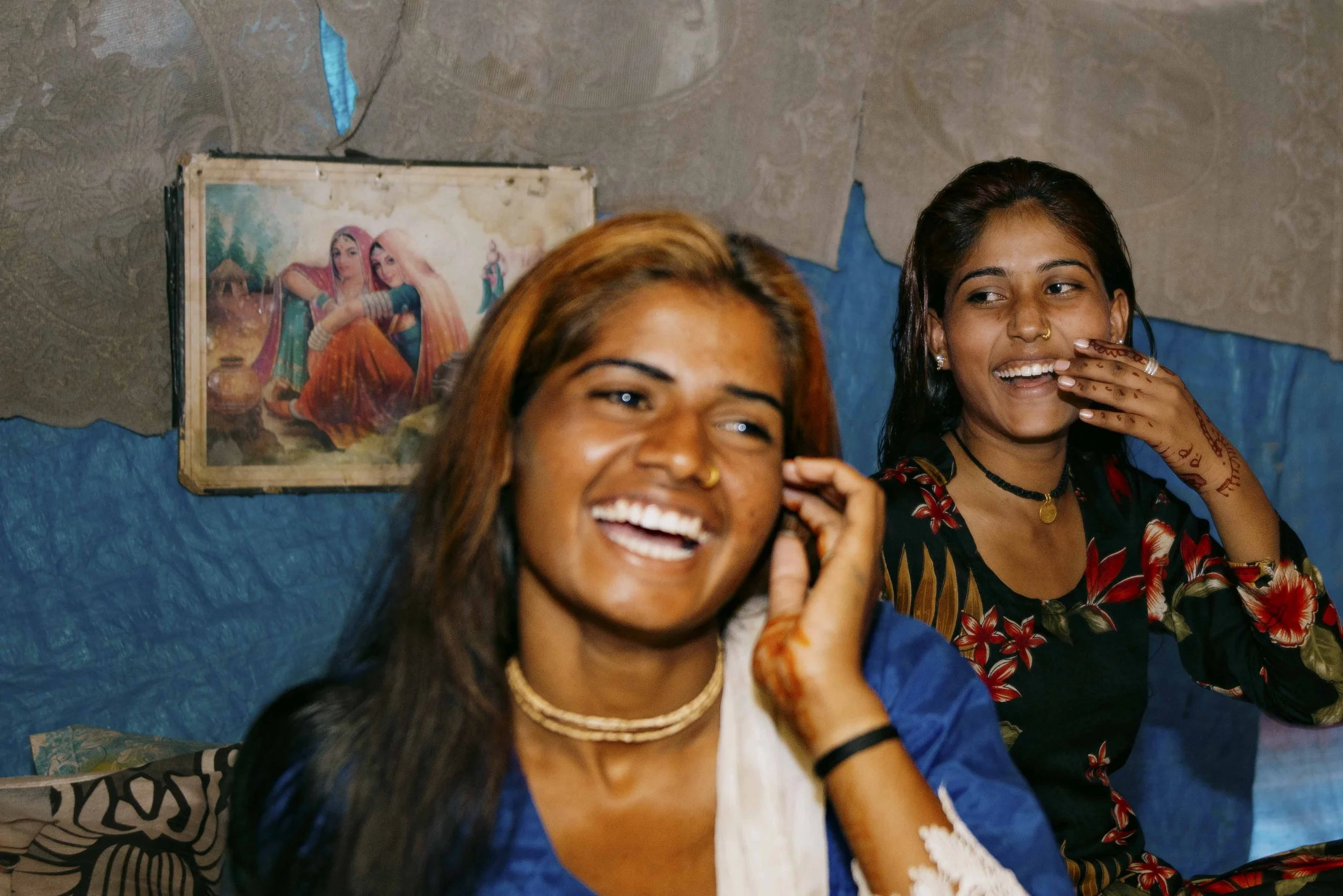 Two young women smiling and laughing, one wearing a blue and white outfit with jewelry, and the other wearing a black floral dress with henna tattoos and jewelry, sitting indoors with a wall featuring a painting of women in traditional Indian attire 