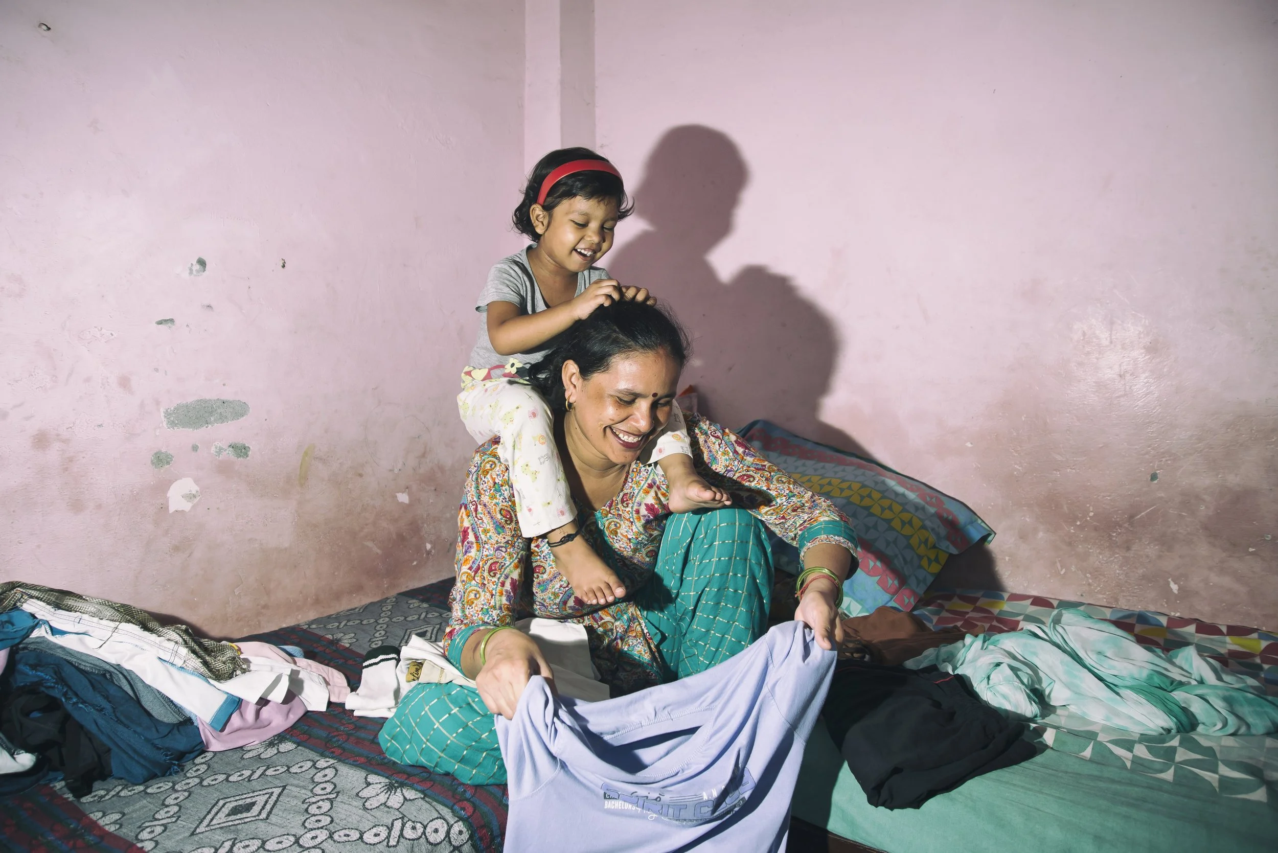 A woman and a young girl on a bed, with the girl playingfully riding on the woman's back, both smiling and enjoying the moment, in a room with pink walls.