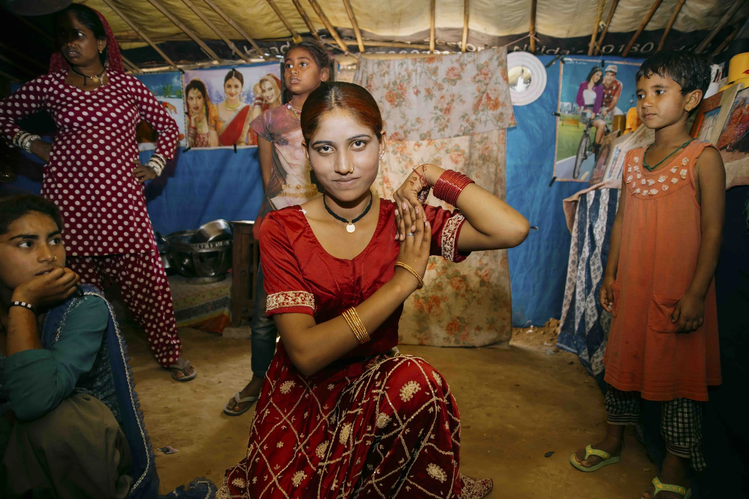 A group of women and children in a tent, with one woman in the center performing a traditional dance or pose, dressed in red and gold attire, surrounded by colorful pictures on the walls.