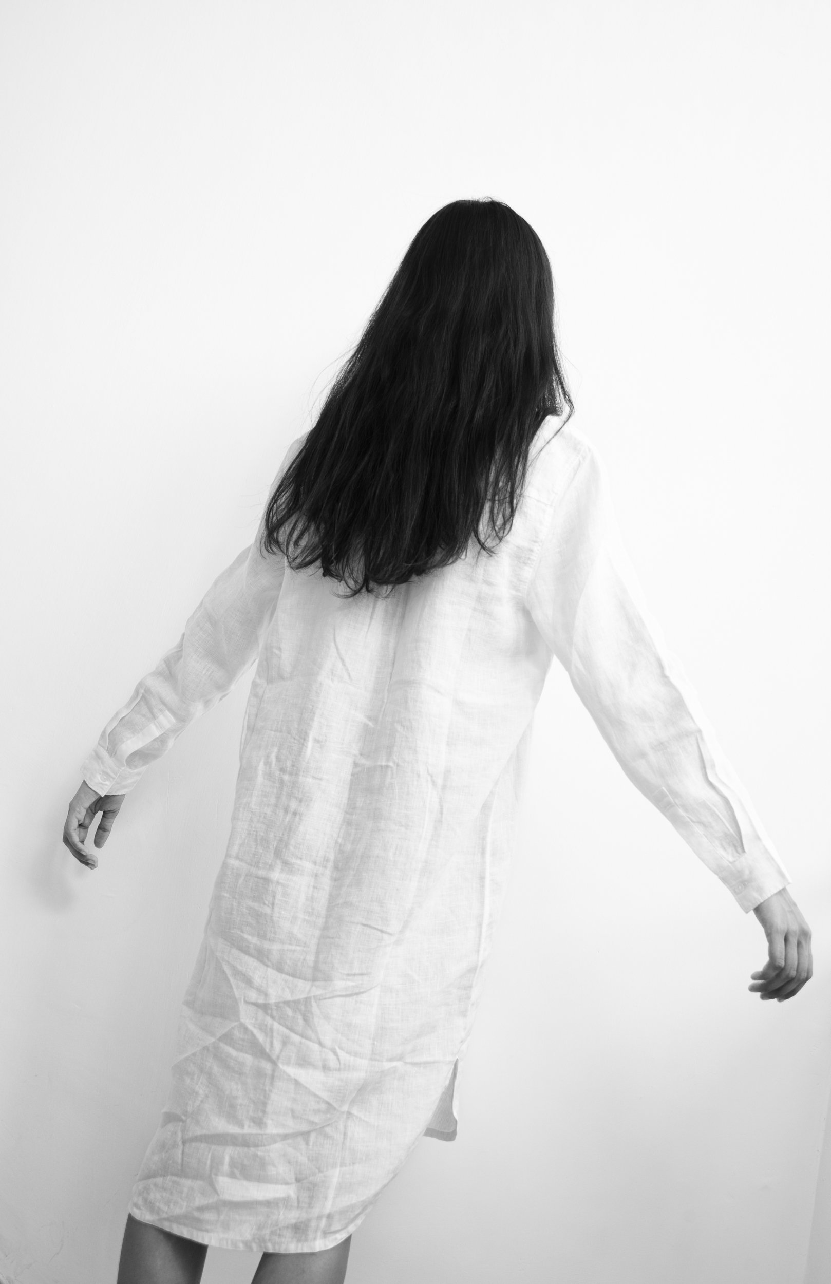 A woman with long dark hair, wearing a light-colored dress, is standing against a plain white background with her arms slightly extended and her head turned away.