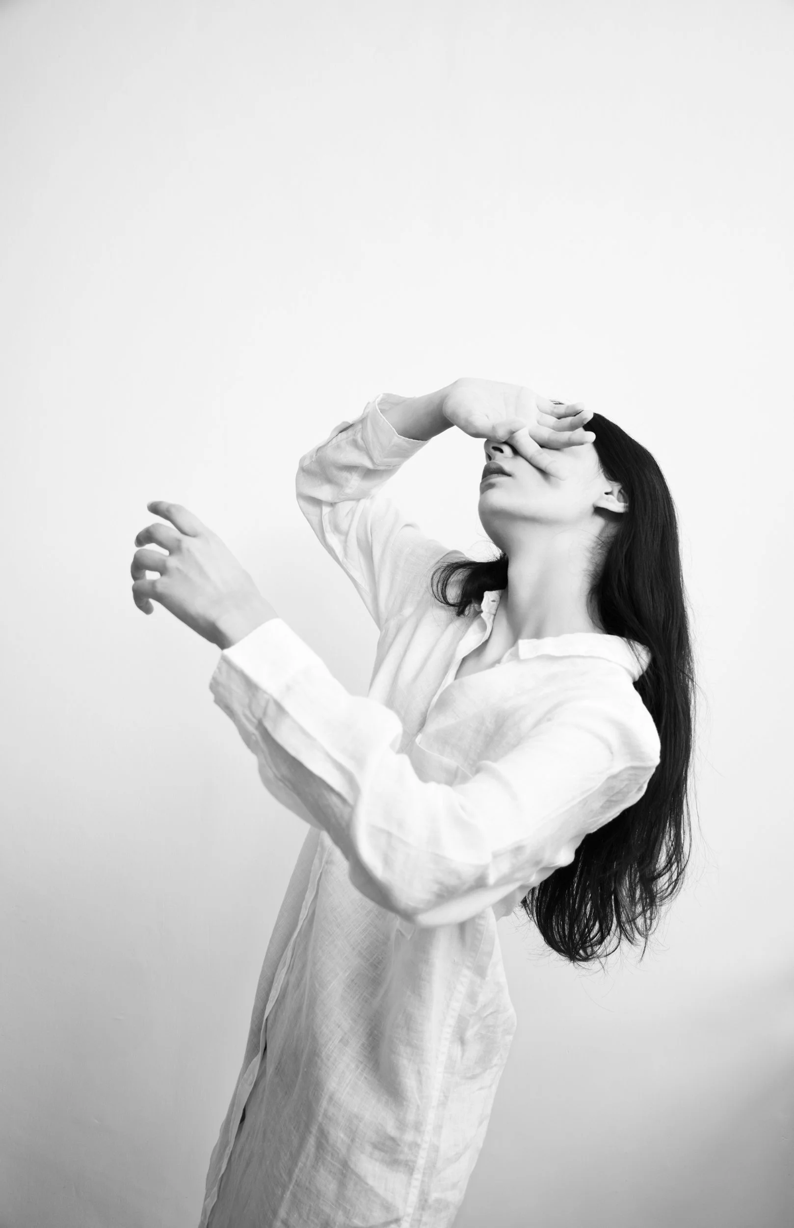 A woman with long dark hair wearing a white shirt, covering her eyes with her left hand, against a plain light background.