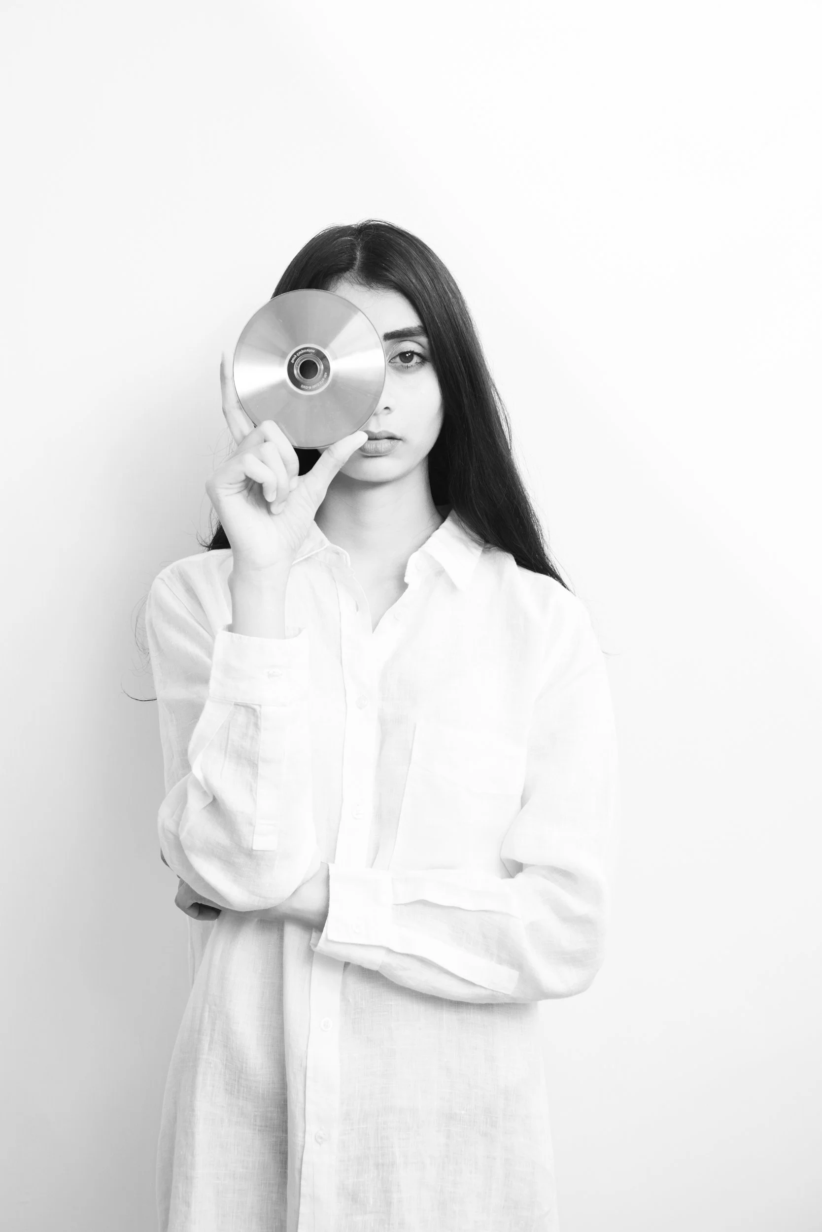 A black and white photo of a woman with long dark hair, wearing a white shirt, holding a CD in front of one eye and looking at the camera.