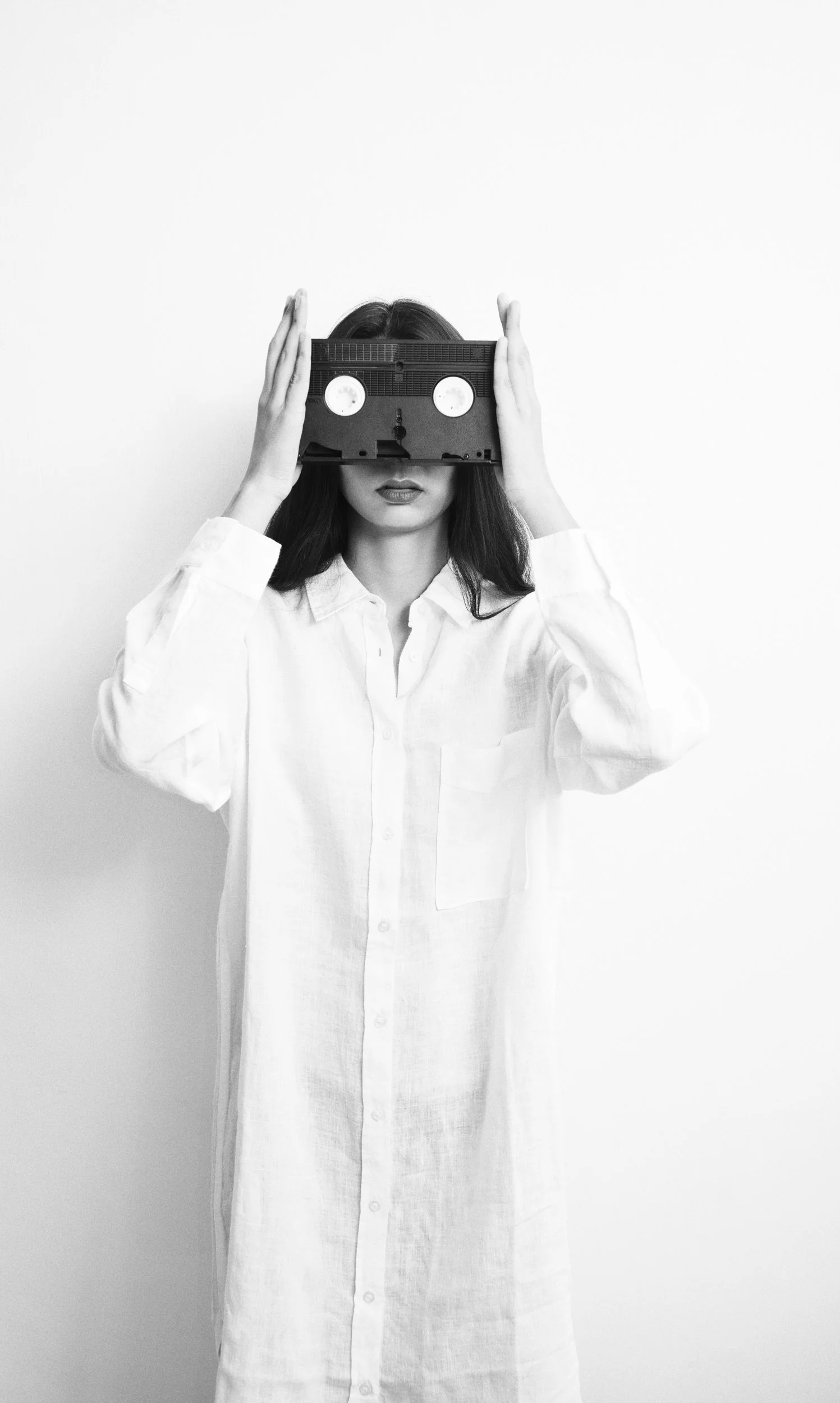 A woman with dark hair wearing a white button-up dress holding a vintage cassette tape over her eyes against a plain wall.