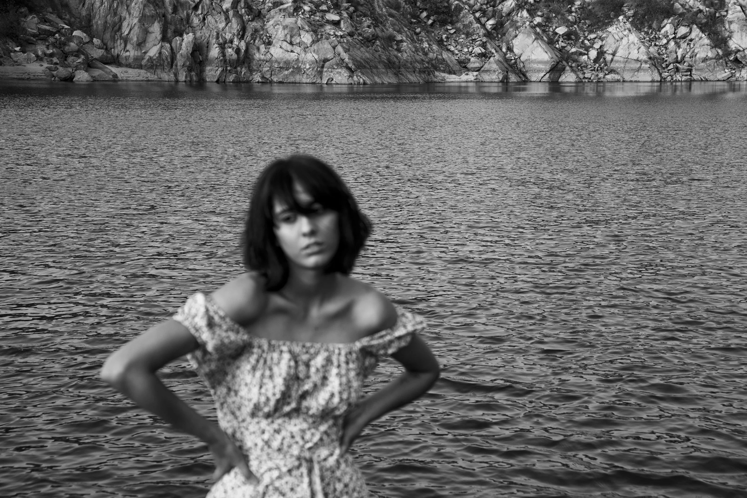 A woman with dark hair and a floral off-shoulder dress standing in front of a body of water with rocky hills in the background.