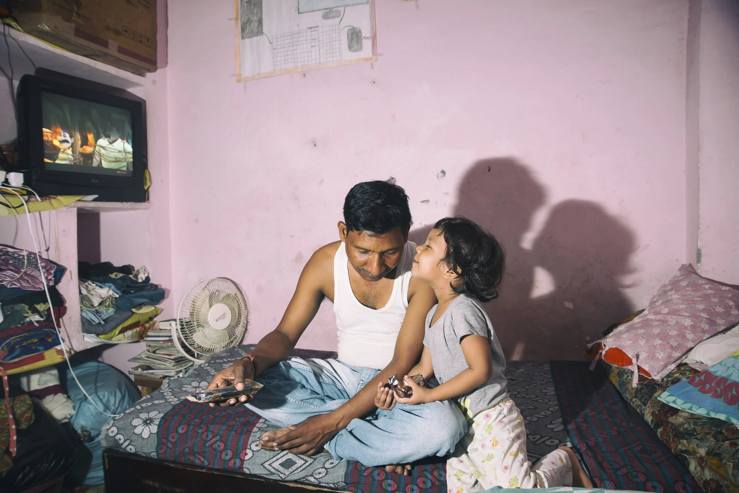 A man and a young girl sitting on a bed in a small, cluttered room, sharing a moment together while looking at a smartphone