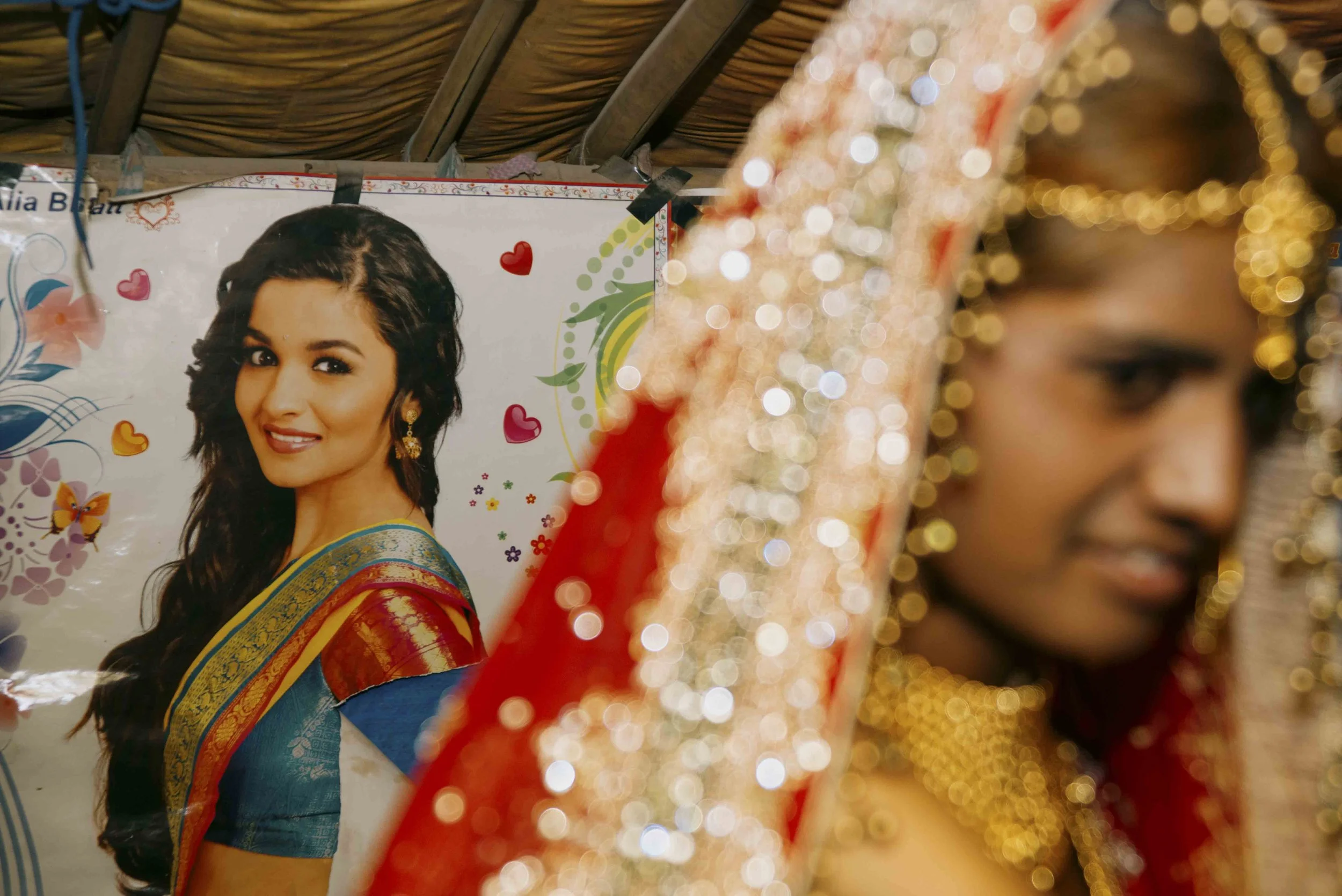 A portrait of a woman with long black hair wearing a colorful, traditional sari with gold embellishments, surrounded by shimmering, ornate fabric in red and gold. A poster of another woman in traditional attire with a blue sari and decorative accesso