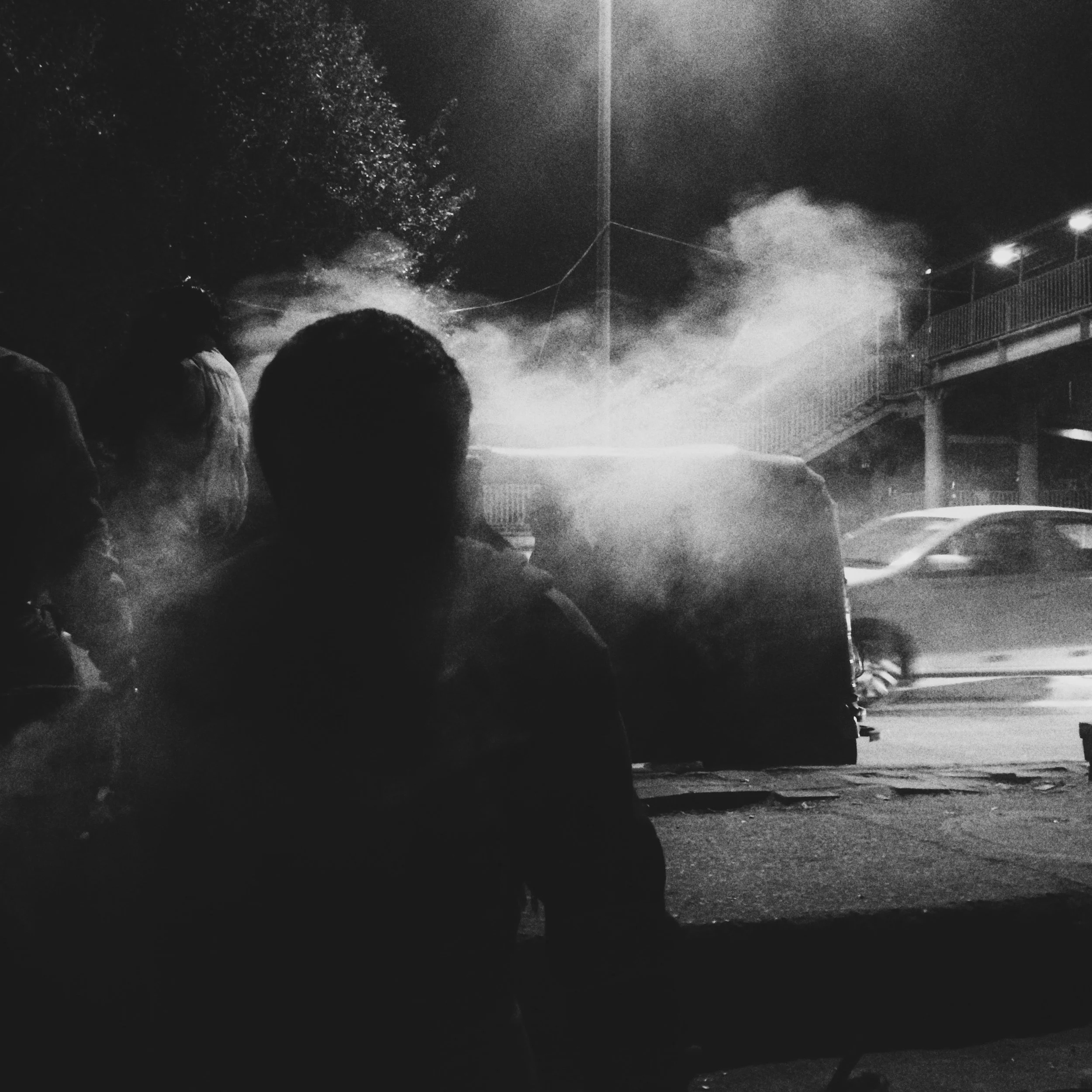 Night scene with people silhouette watching a street fire or explosion with smoke, cars, and a staircase in the background.